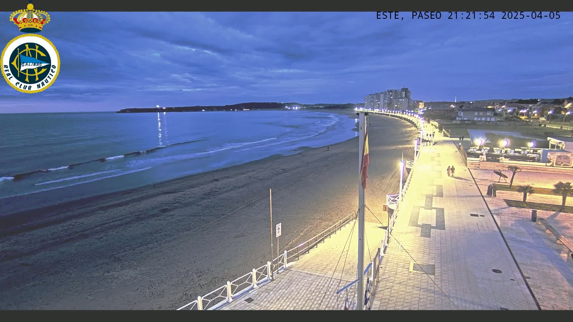 A beach at twilight with calm waves, a boardwalk lined with lights, and a few people walking along the shore under a partly cloudy sky.