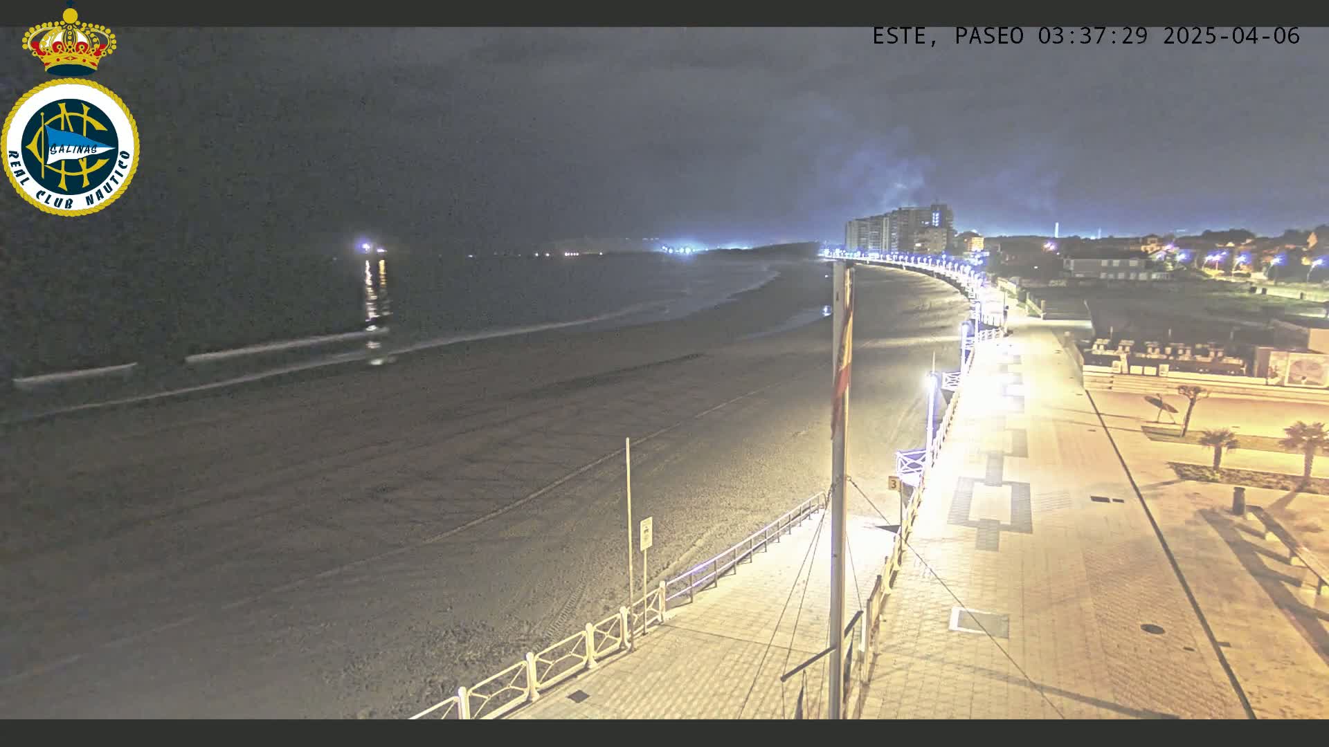 A nighttime, overhead view shows a mostly empty, sandy beach next to a well-lit boardwalk that curves alongside buildings.