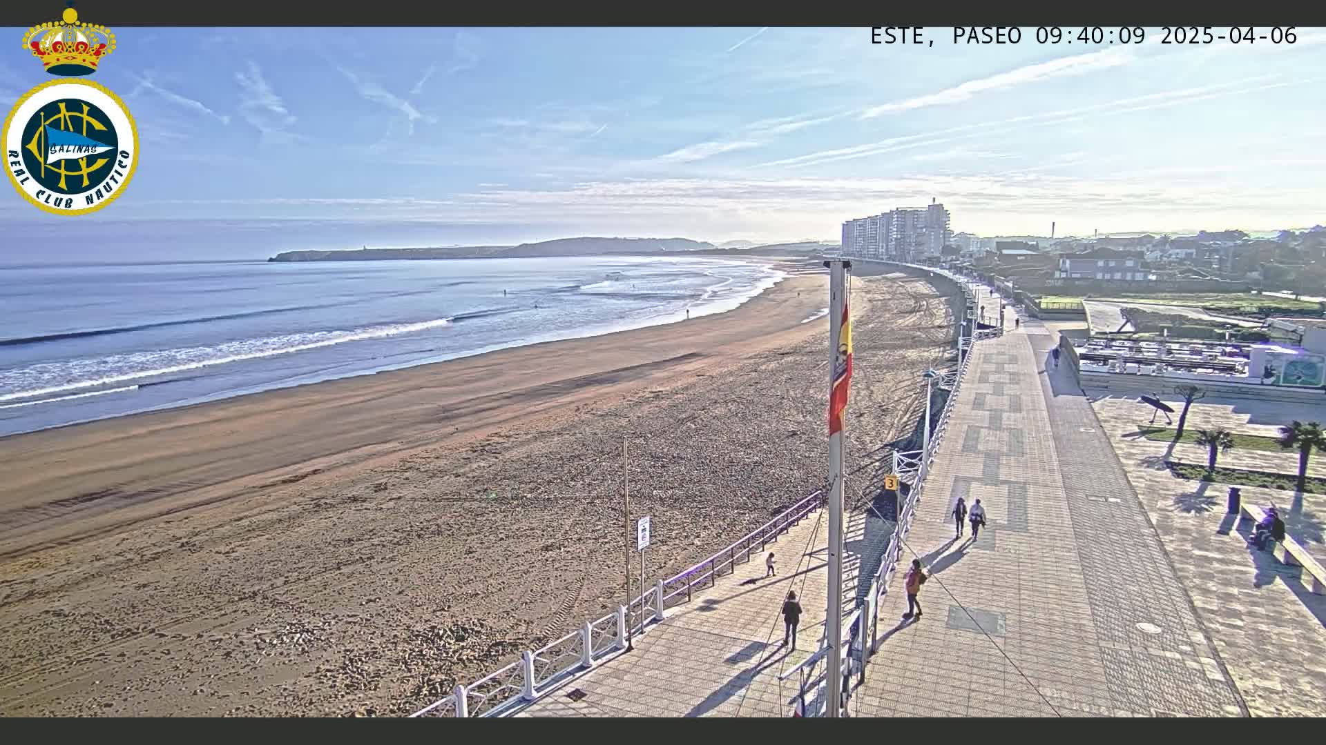 A sandy beach with gentle waves under a clear, sunny sky is bordered by a paved walkway with a few people walking, and buildings are visible in the distance.