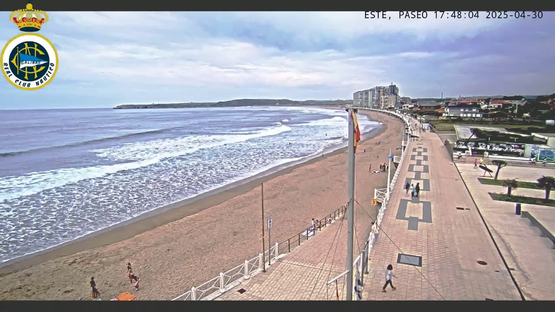 A sandy beach with moderate waves under a partly cloudy sky is bordered by a paved walkway and buildings.