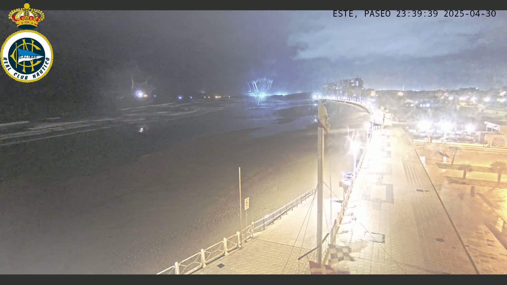 A nighttime view of a beach at low tide with a boardwalk and buildings in the distance under a cloudy sky.