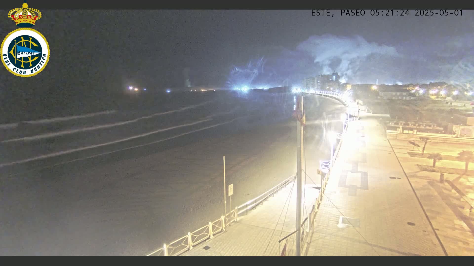 A nighttime view of a beach with gentle waves, a paved boardwalk with lights, and buildings in the background under a cloudy sky.