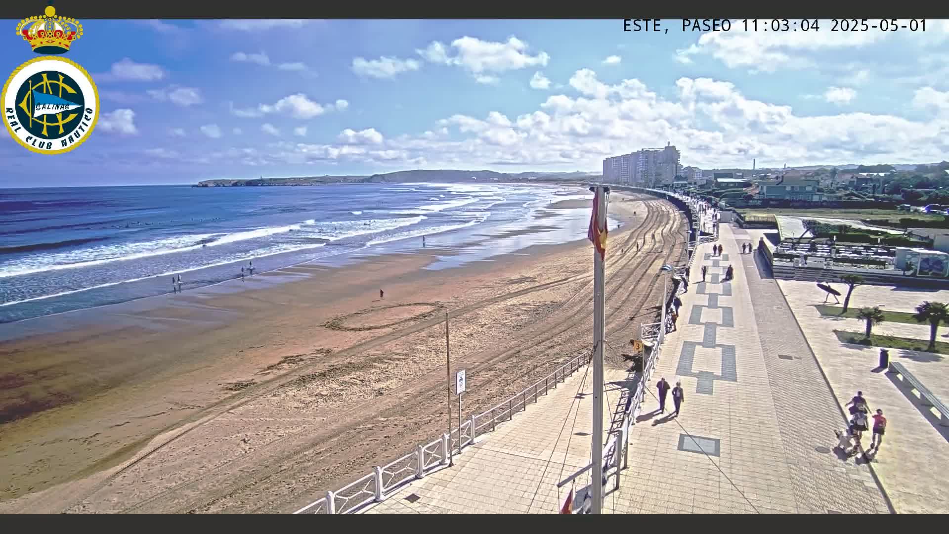 A sandy beach with small waves under a partly cloudy sky is bordered by a paved walkway and buildings.