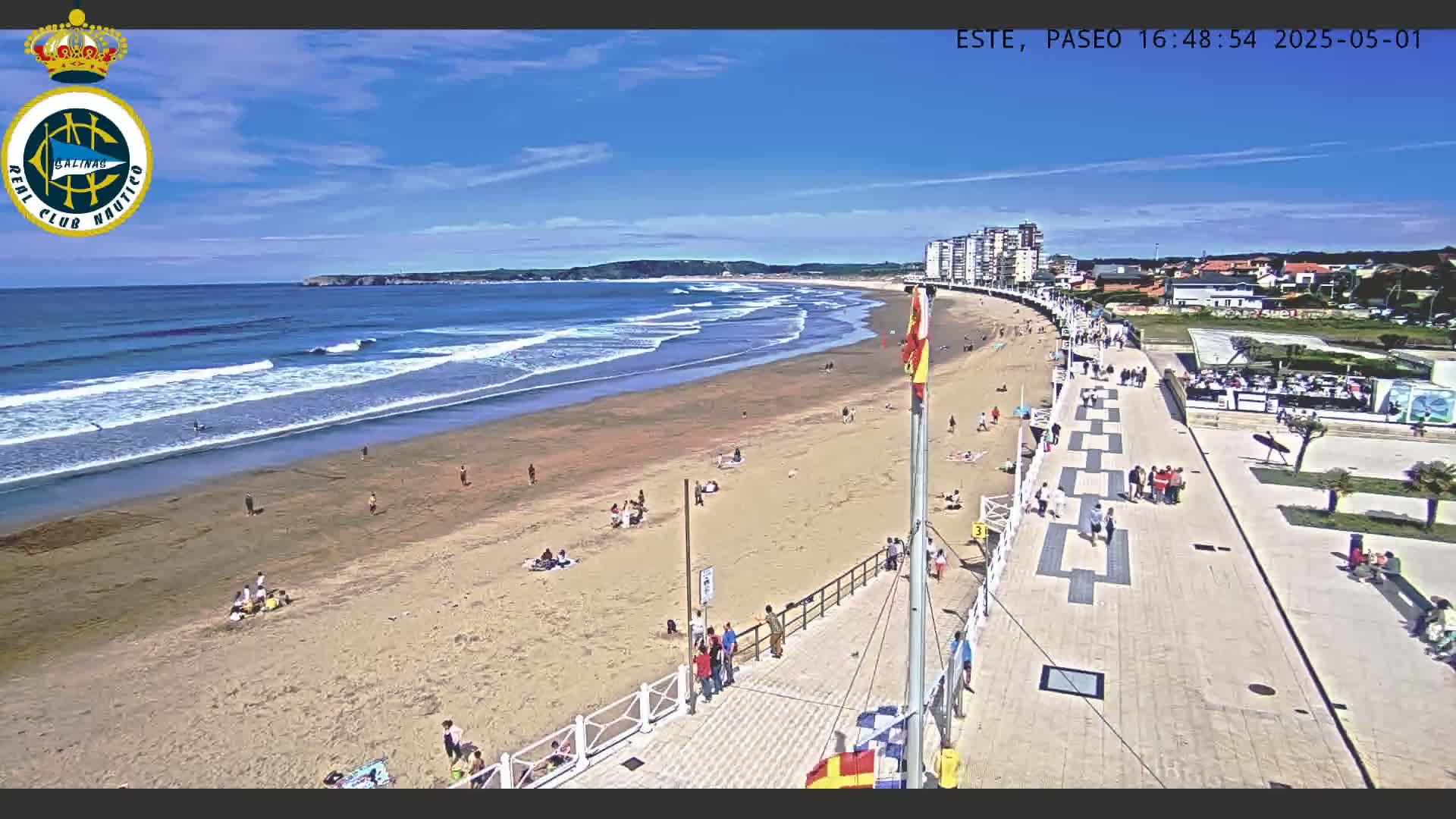 A sandy beach with moderate waves under a clear blue sky is bordered by a paved promenade lined with people and buildings.