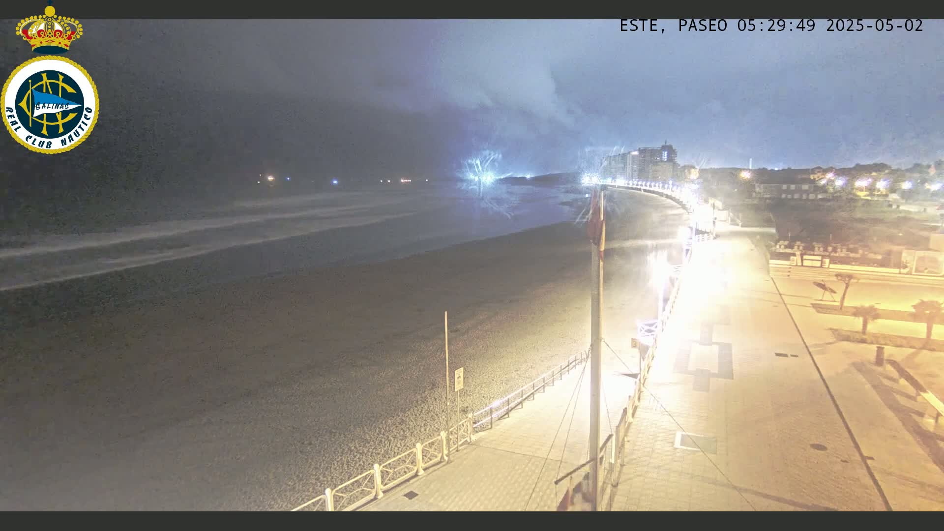 A nighttime view of a dark, sandy beach with gentle waves under a cloudy sky, a brightly lit promenade stretches alongside it, leading to buildings in the distance.