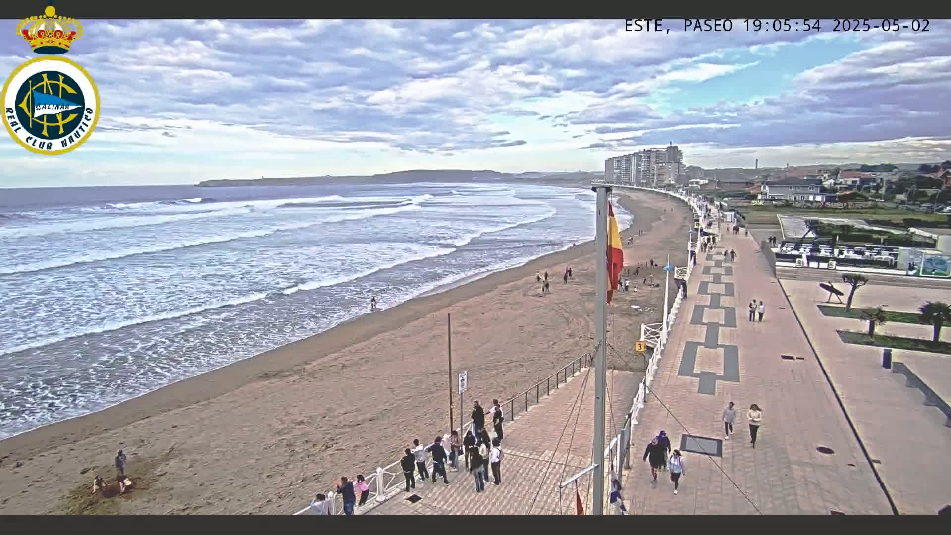 A sandy beach with moderate waves under a partly cloudy sky is shown, with people walking on the adjacent boardwalk and beach.