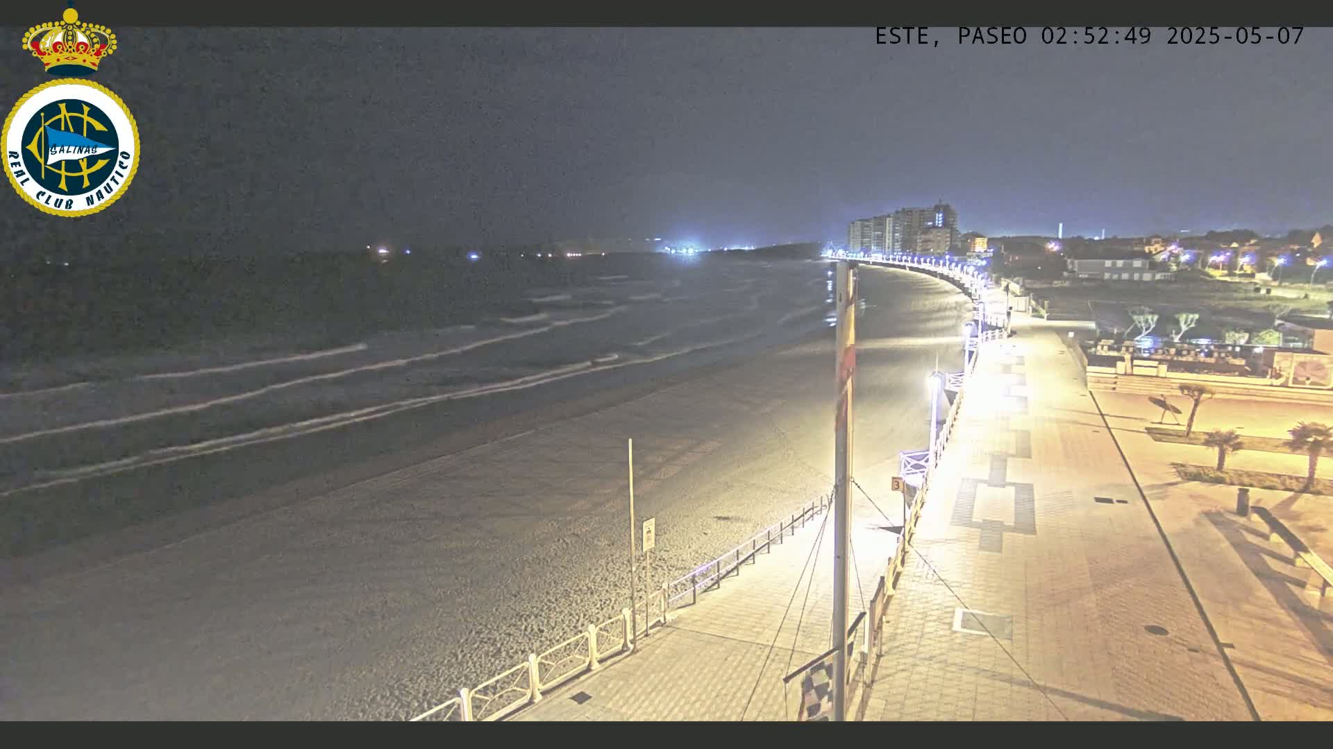 A nighttime view of a beach with gentle waves, a paved walkway lined with lights, and buildings in the background.