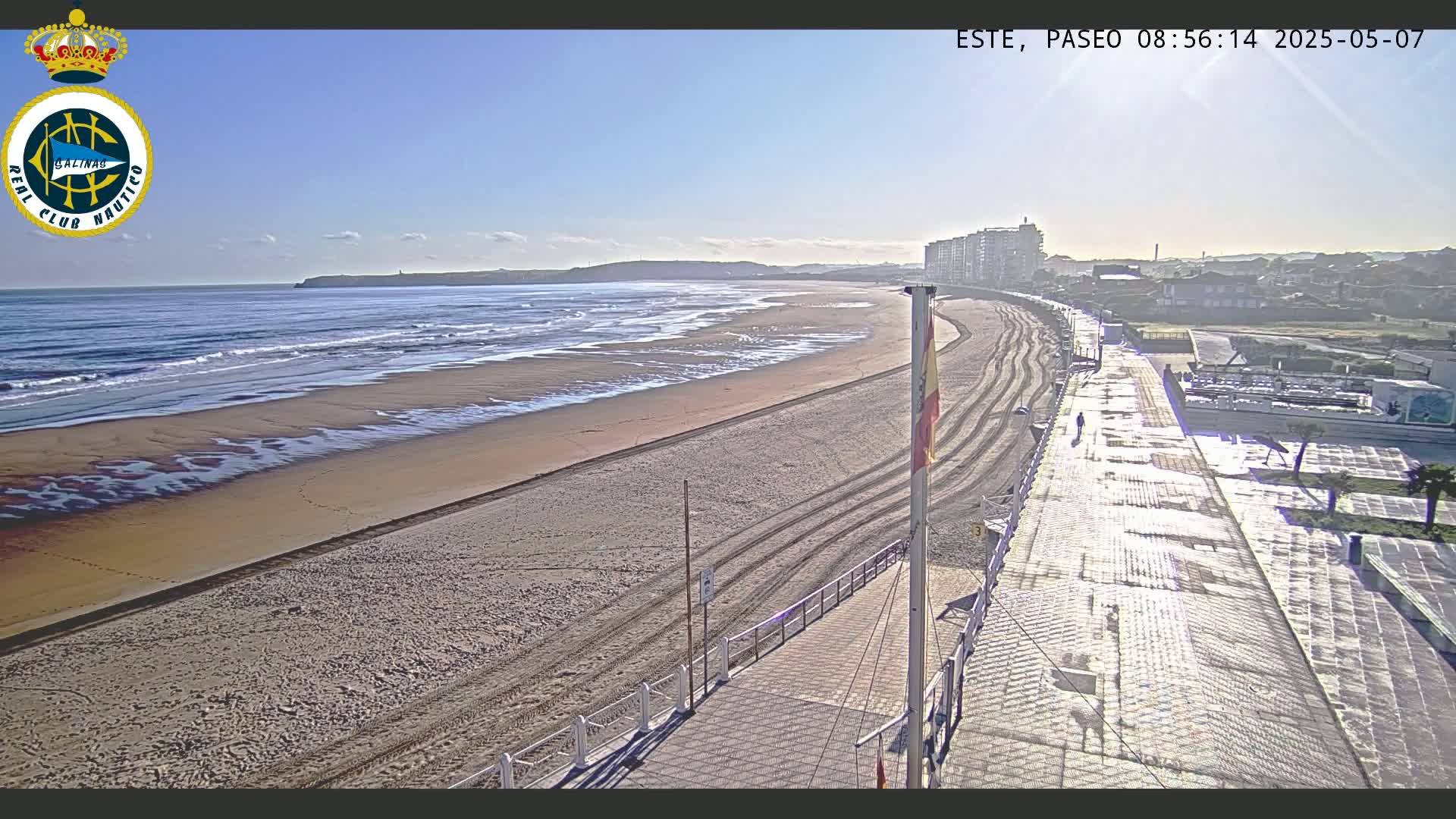 A sandy beach with gentle waves stretches along a paved boardwalk under a sunny sky, with buildings visible in the distance.