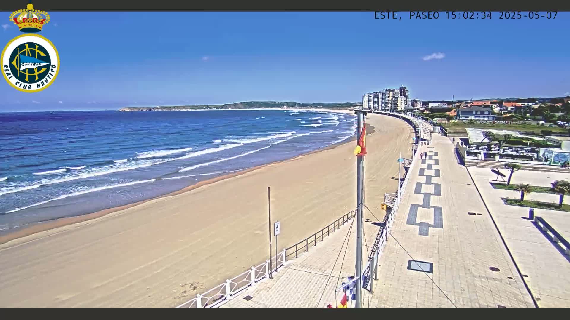 A sandy beach with gentle waves under a clear blue sky is bordered by a paved walkway lined with buildings, leading to a town.