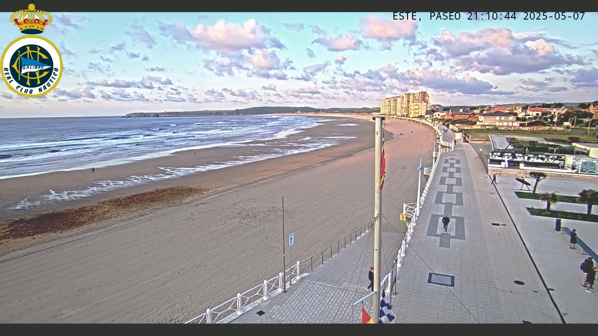 A long sandy beach with gentle waves under a partly cloudy sky is bordered by a paved walkway and buildings.