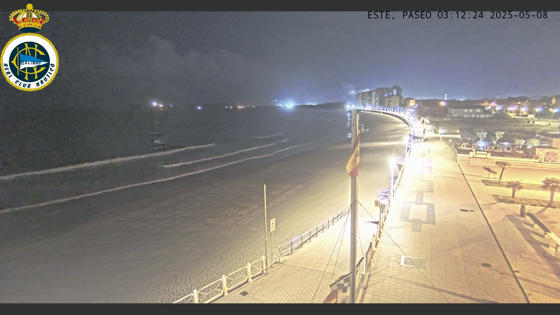 A nighttime view of a mostly empty beach with gently lapping waves, a paved walkway with lights, and buildings in the distance under a partly cloudy sky.