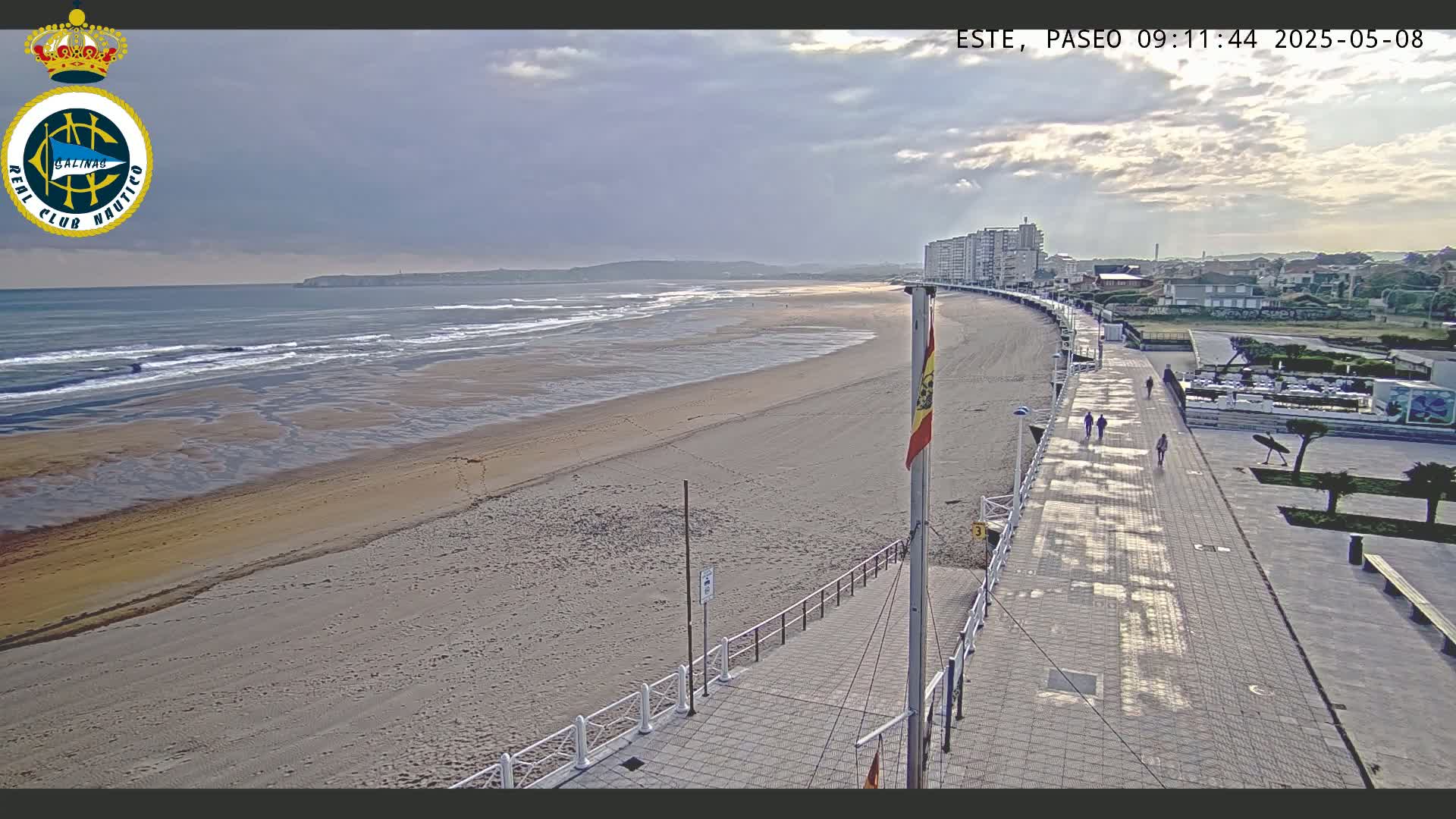 A sandy beach with gentle waves under a partly cloudy sky is viewed from a boardwalk next to buildings.