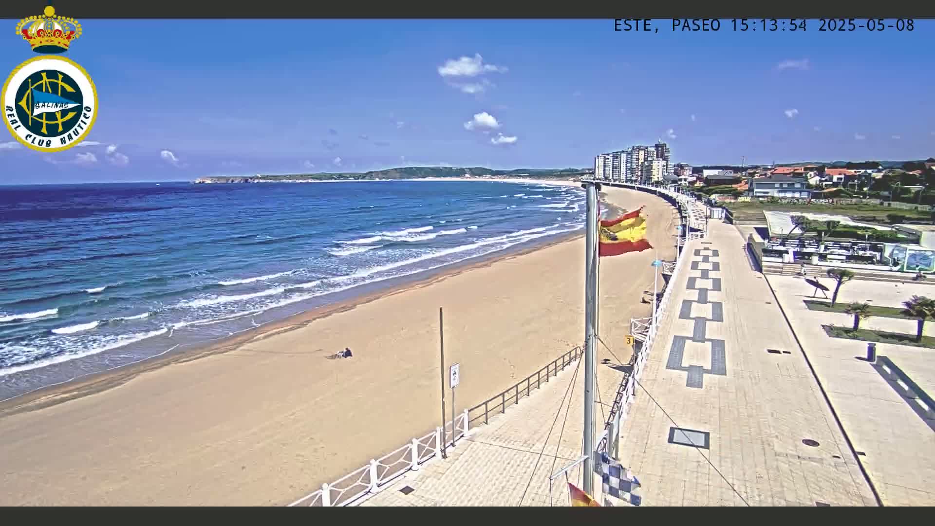 A sandy beach with gentle waves under a clear blue sky is bordered by a paved walkway leading to buildings, with a Spanish flag waving nearby.