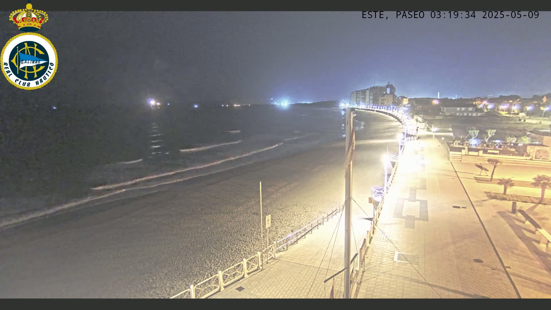 A dark, nighttime beach scene shows gently rolling waves on a sandy shore next to a paved walkway lined with lights, leading to buildings in the distance.