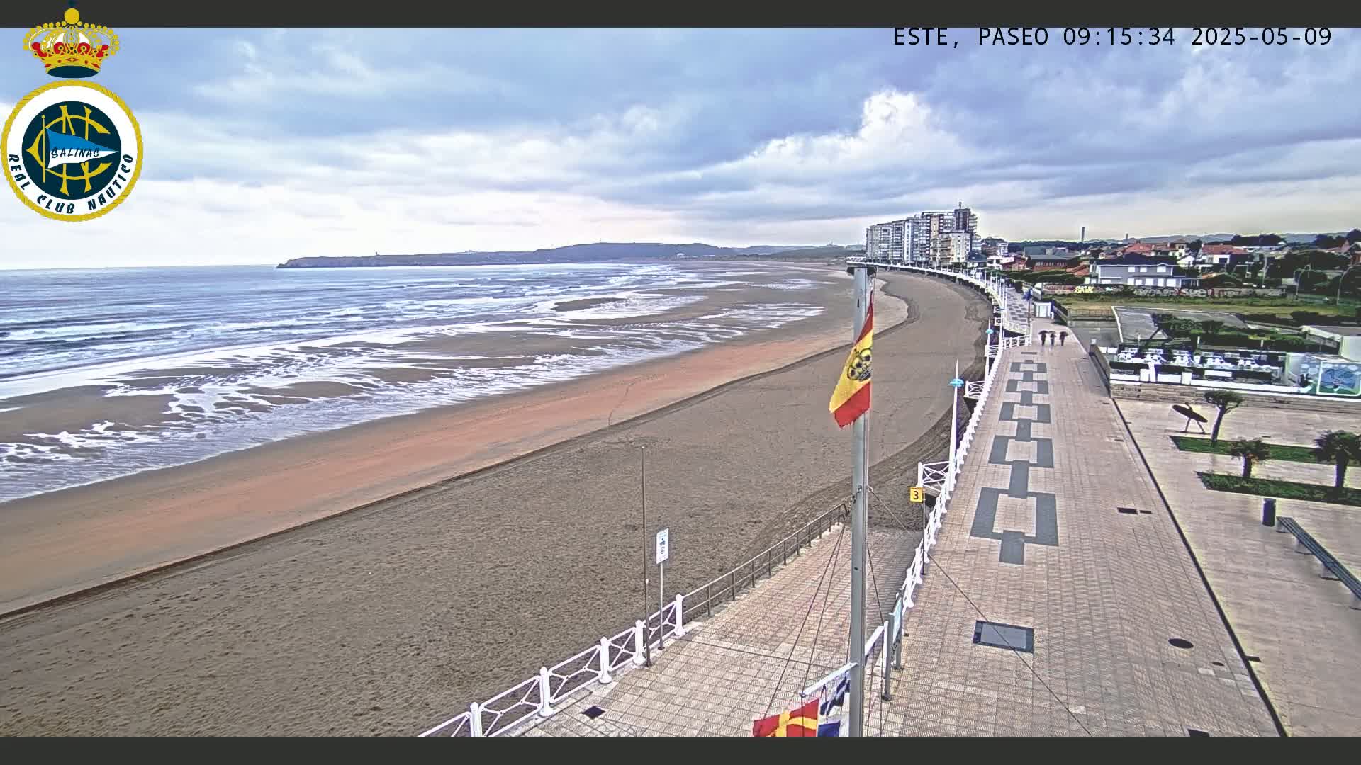 A sandy beach with gentle waves under a cloudy sky is bordered by a paved walkway and buildings, with a Spanish flag visible.