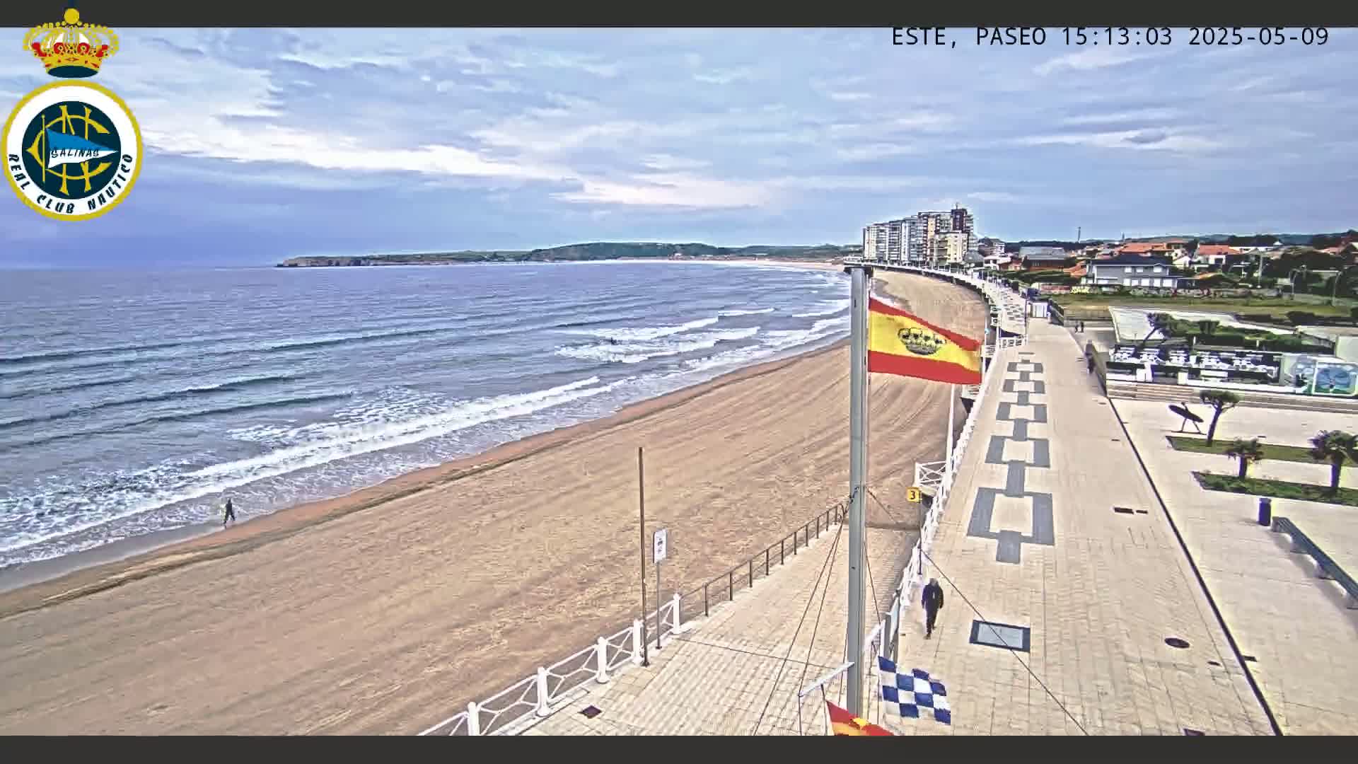 A sandy beach with small waves stretches beneath a partly cloudy sky next to a paved promenade lined with buildings.