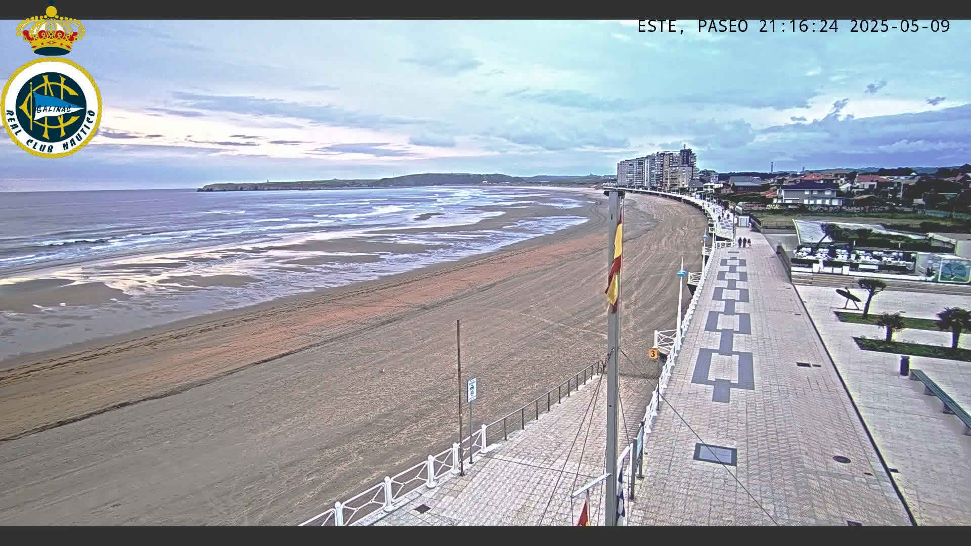 A long sandy beach with gentle waves under a partly cloudy sky is bordered by a paved walkway and buildings.