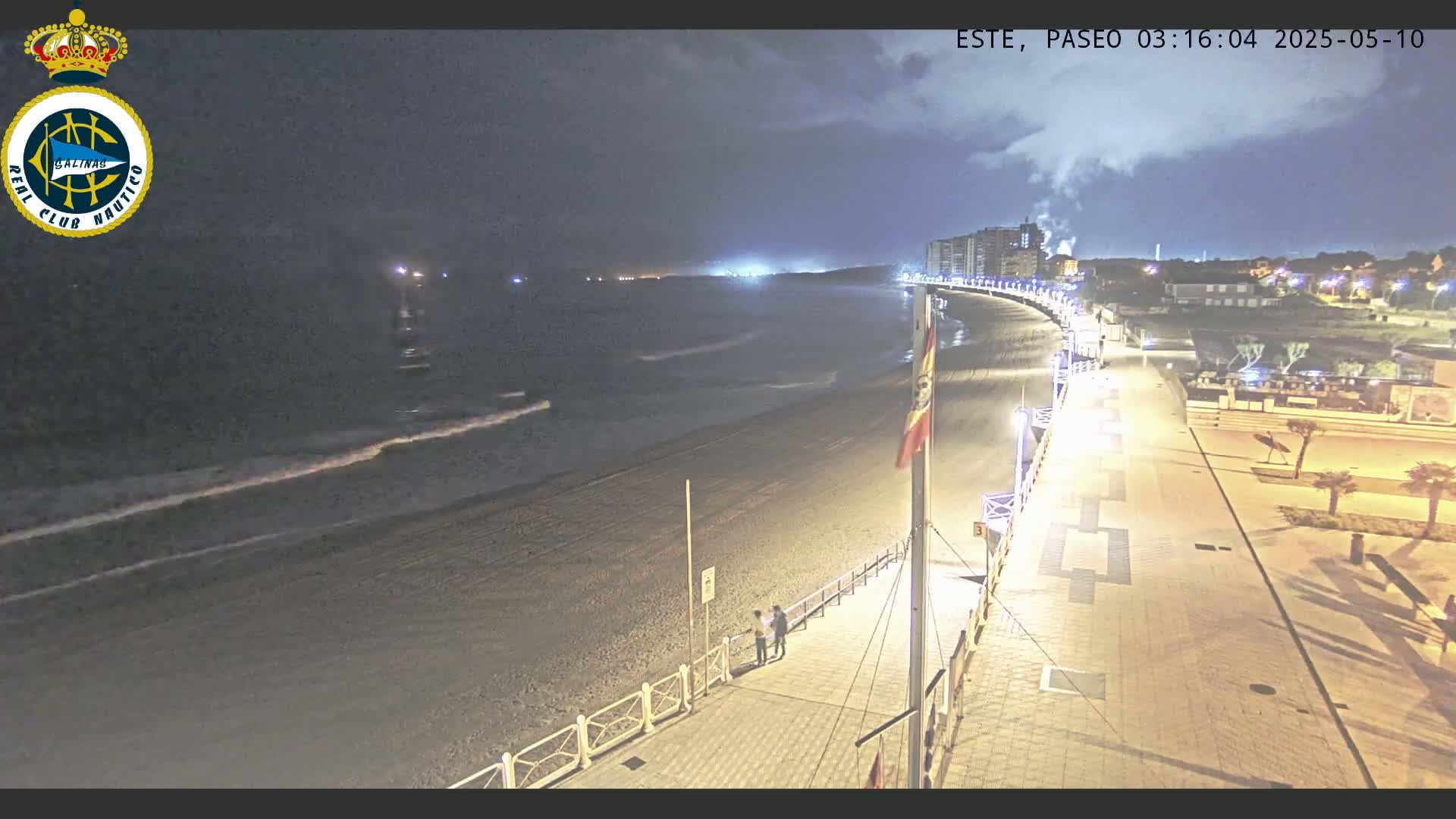 A nighttime view of a beach with gentle waves, a paved boardwalk with lights, and buildings in the distance under a partly cloudy sky.