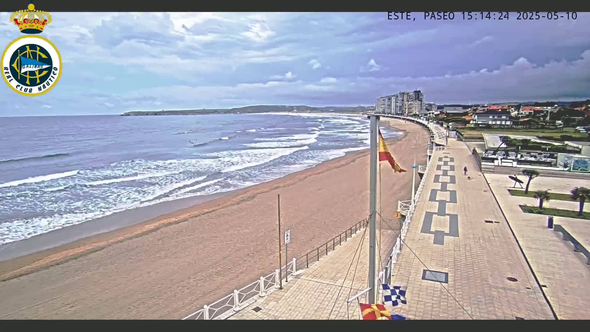 A sandy beach with moderate waves under a partly cloudy sky is adjacent to a paved walkway and buildings.
