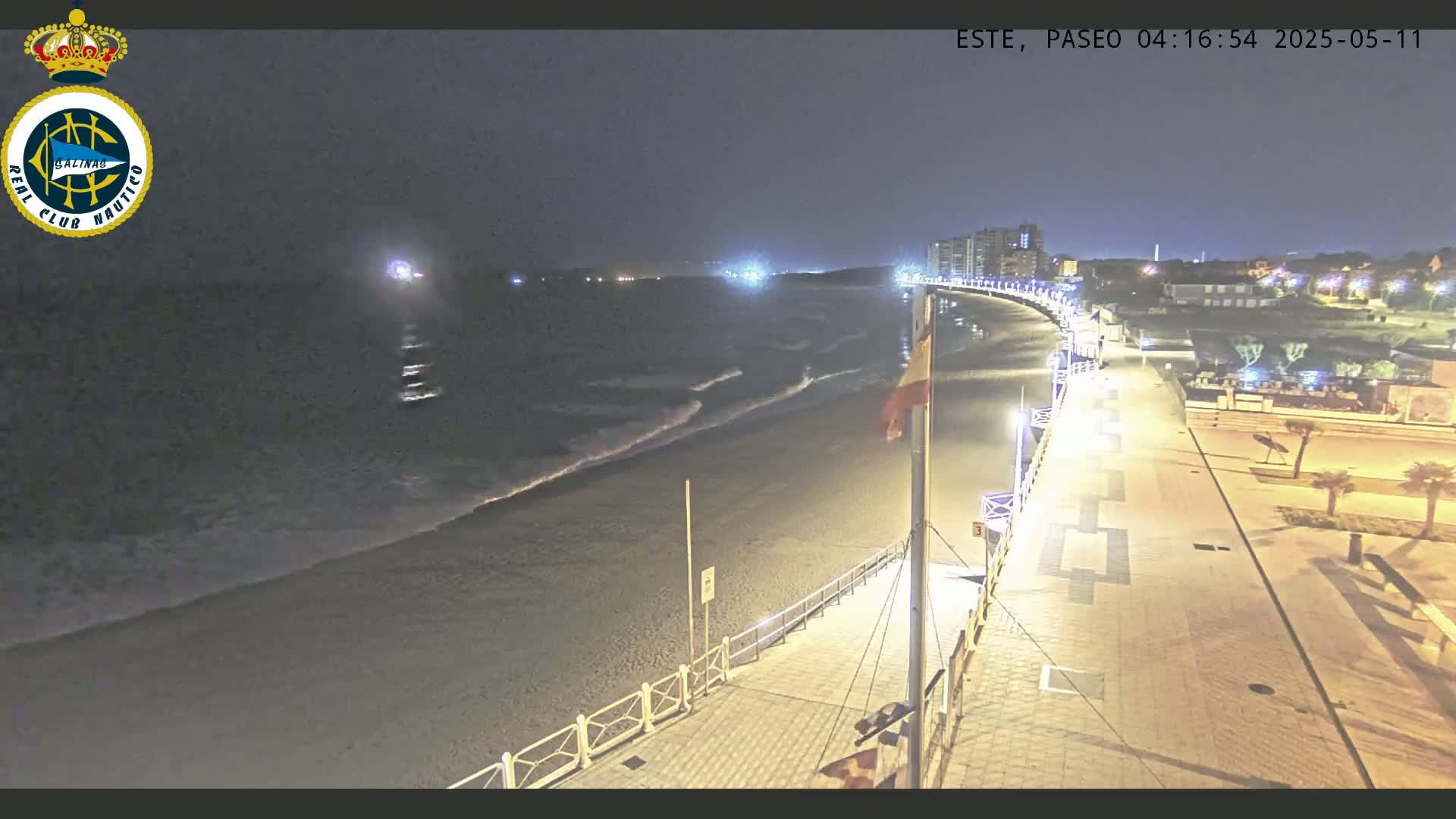 A nighttime view shows a sandy beach with gentle waves lapping the shore, a paved boardwalk with lights, and buildings in the distance.