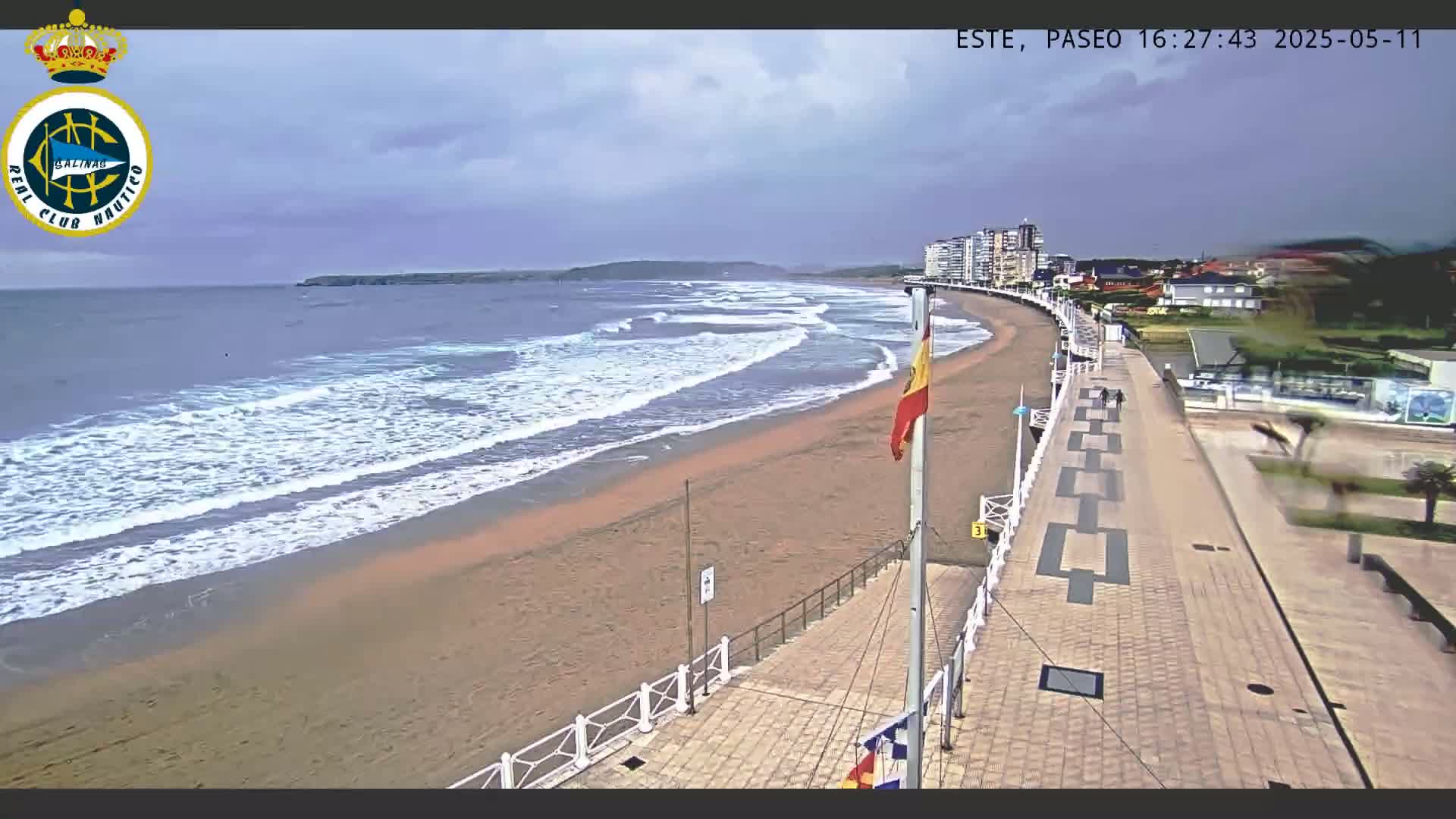 A sandy beach with moderate waves under an overcast sky is bordered by a paved walkway leading to buildings, with a Spanish flag visible.
