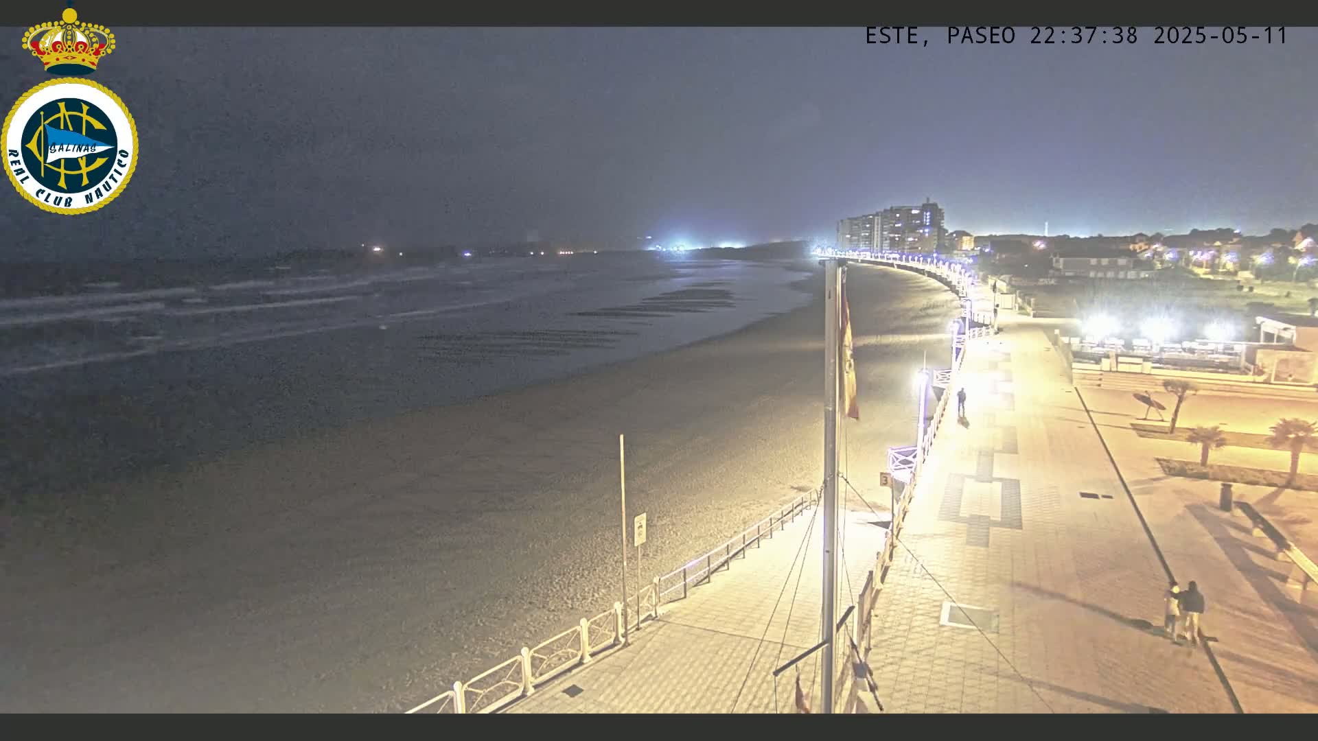 A nighttime view of a mostly empty beach with a paved walkway and buildings in the distance, under a dark, cloudy sky with gentle waves.
