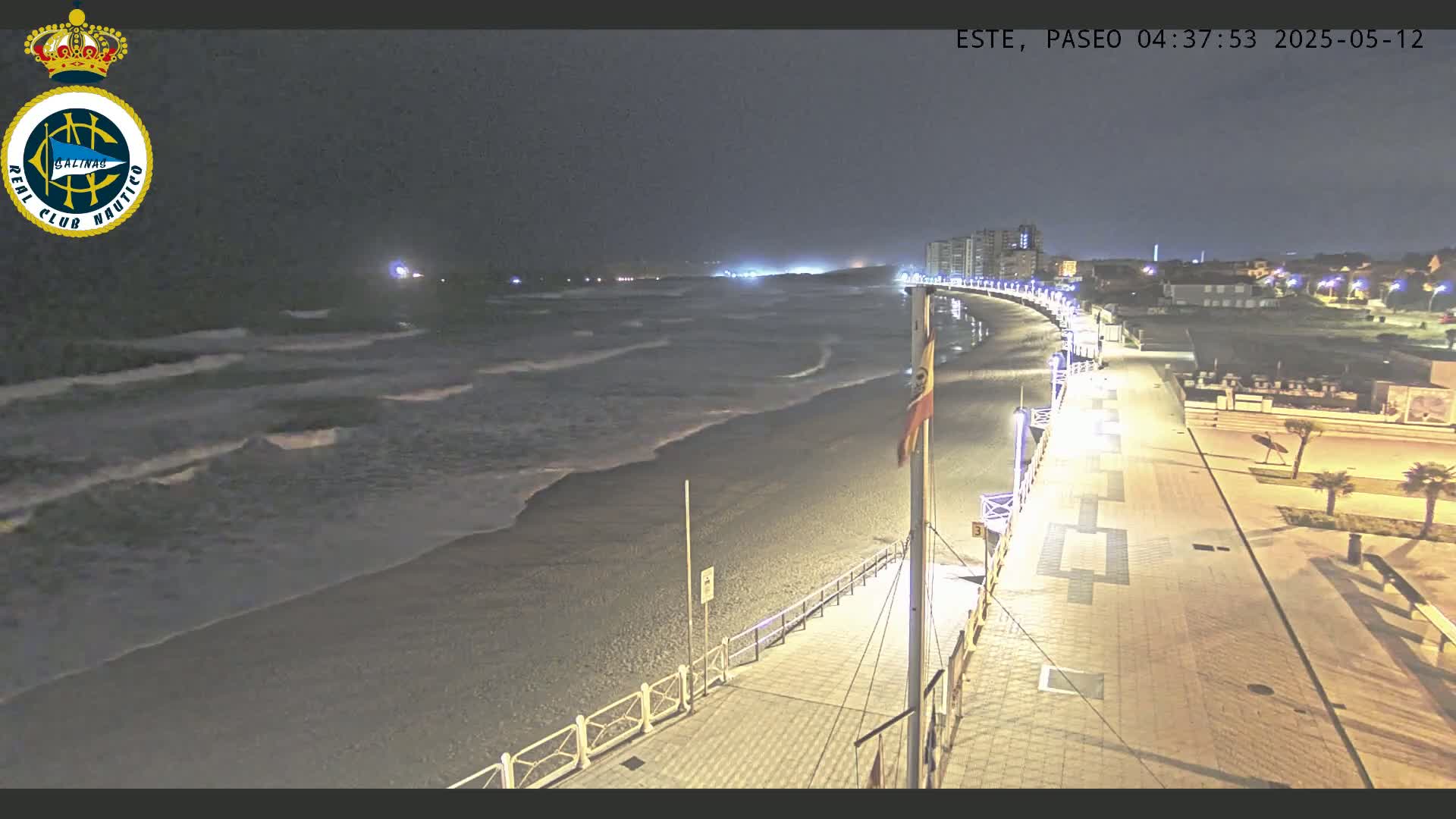 A nighttime view of a beach with gentle waves lapping the shore, a paved boardwalk lined with lights, and buildings in the distance.
