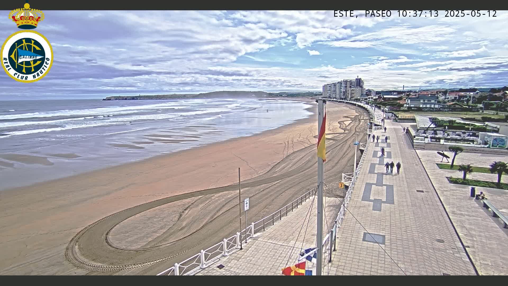 A sandy beach with small waves under a partly cloudy sky is viewed from a paved boardwalk that leads to buildings along the coast.