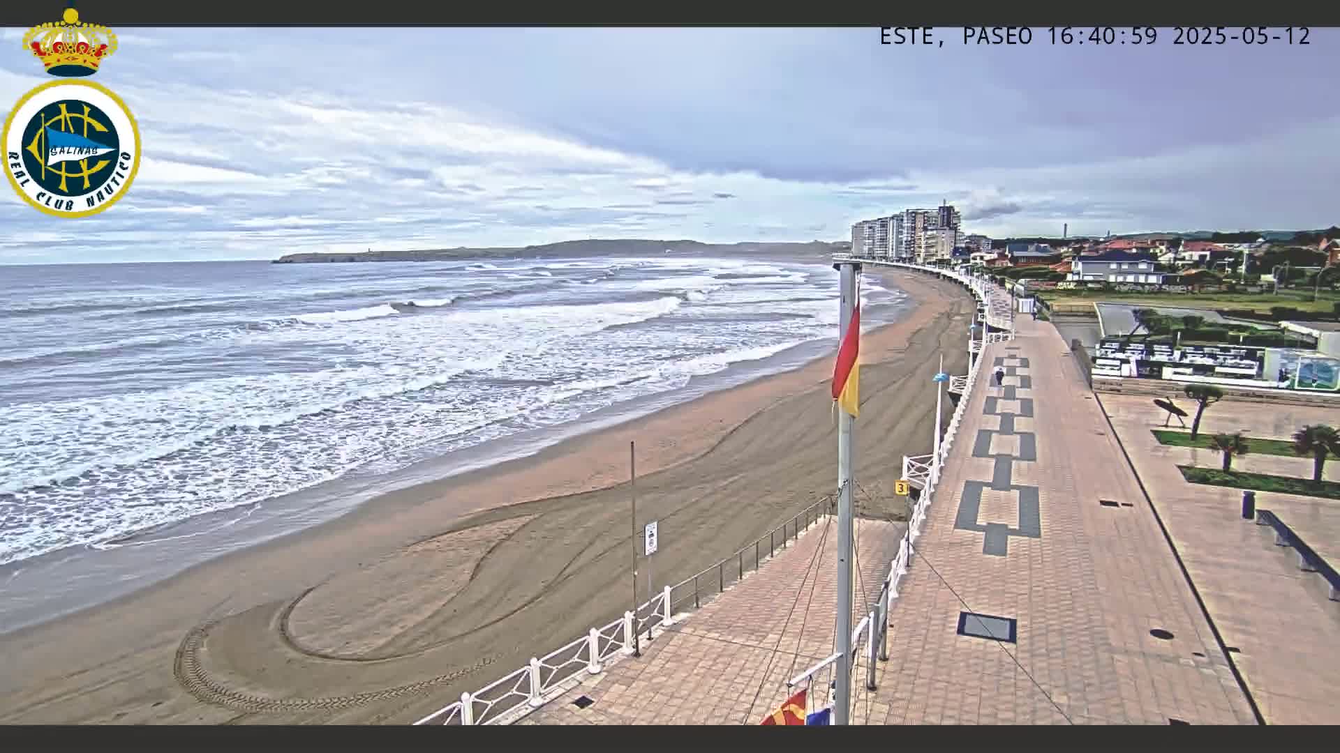 A sandy beach with moderate waves under an overcast sky is bordered by a paved walkway and buildings.