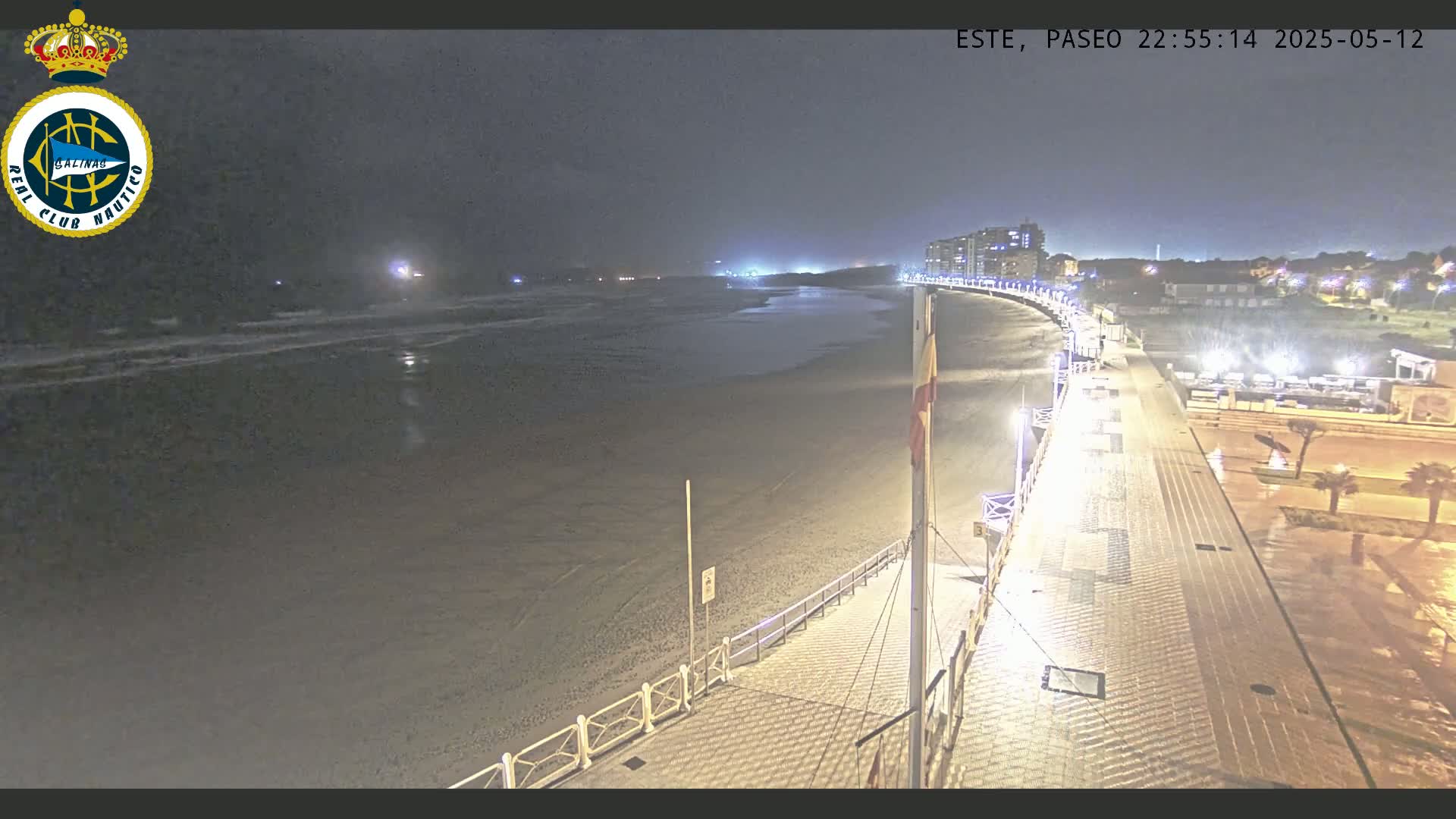 A nighttime view of a mostly empty beach and boardwalk, with calm waves and distant lights from buildings.