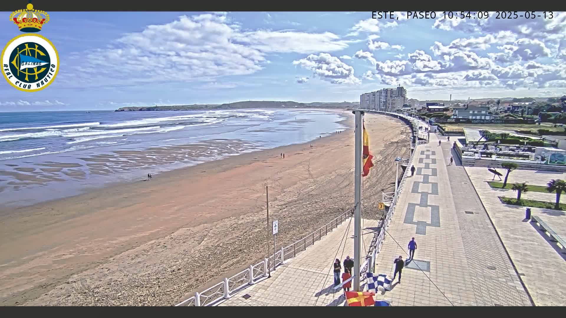 A sandy beach with gentle waves under a partly cloudy sky is bordered by a paved walkway with several people walking along it, and buildings are visible in the background.