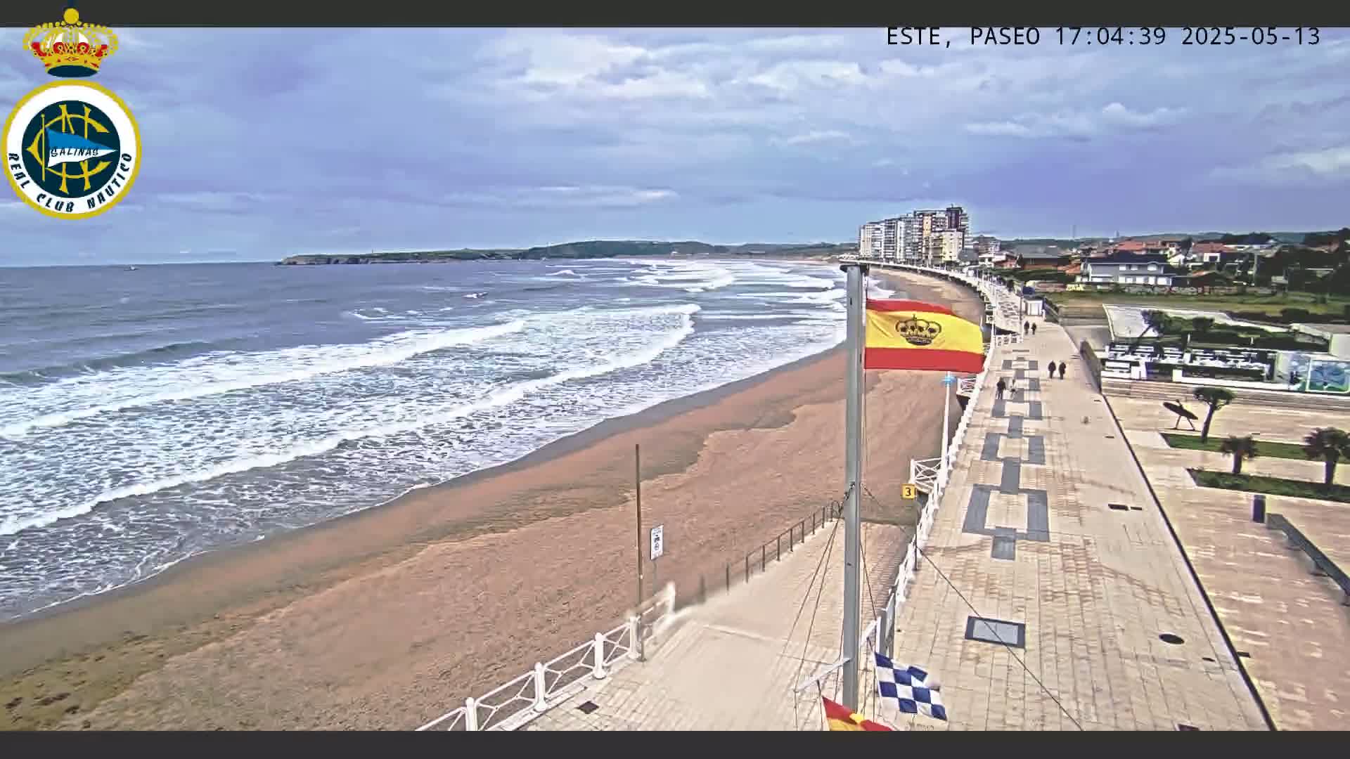 A sandy beach with gentle waves under a partly cloudy sky is bordered by a paved walkway with several pedestrians, and buildings are visible in the background.