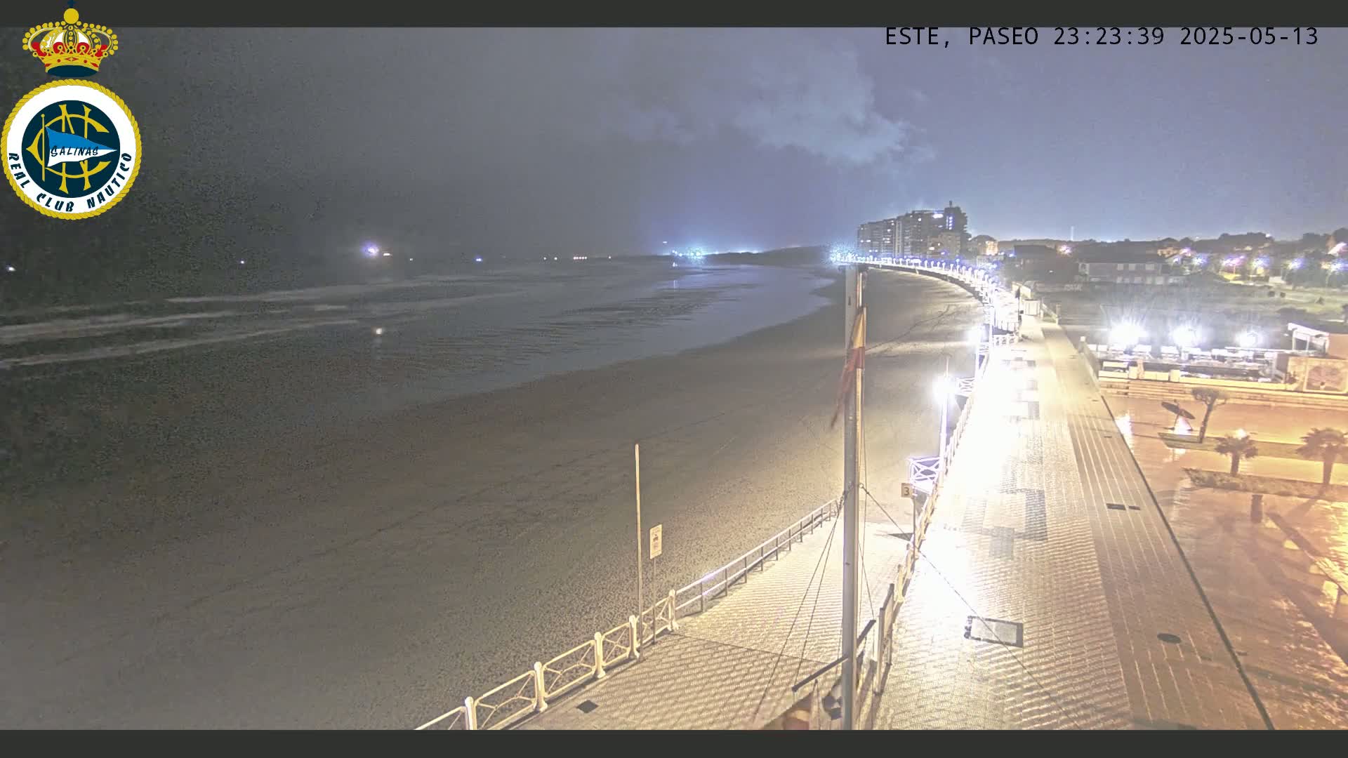 A nighttime view of a beach boardwalk and shoreline under a cloudy sky, with calm waves lapping the shore.