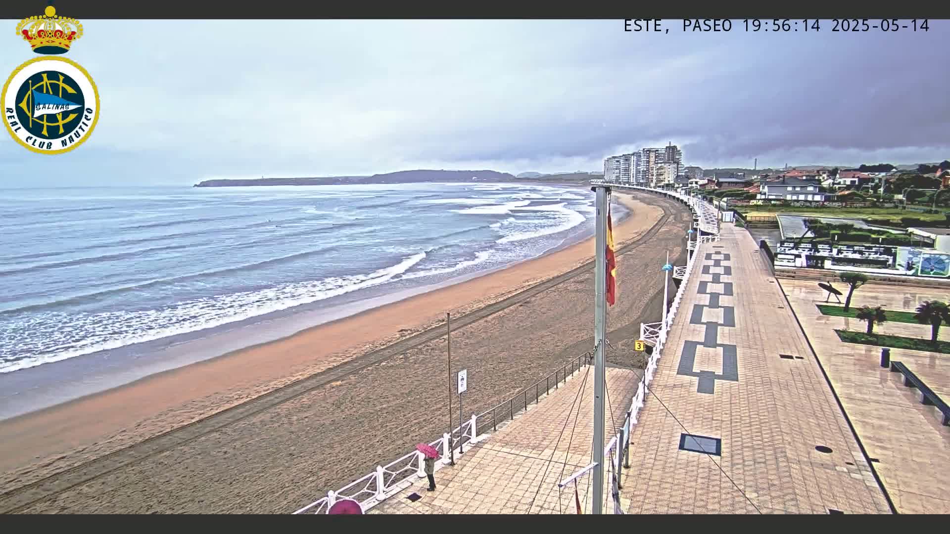 A sandy beach with gentle waves under an overcast sky is bordered by a paved boardwalk and buildings.