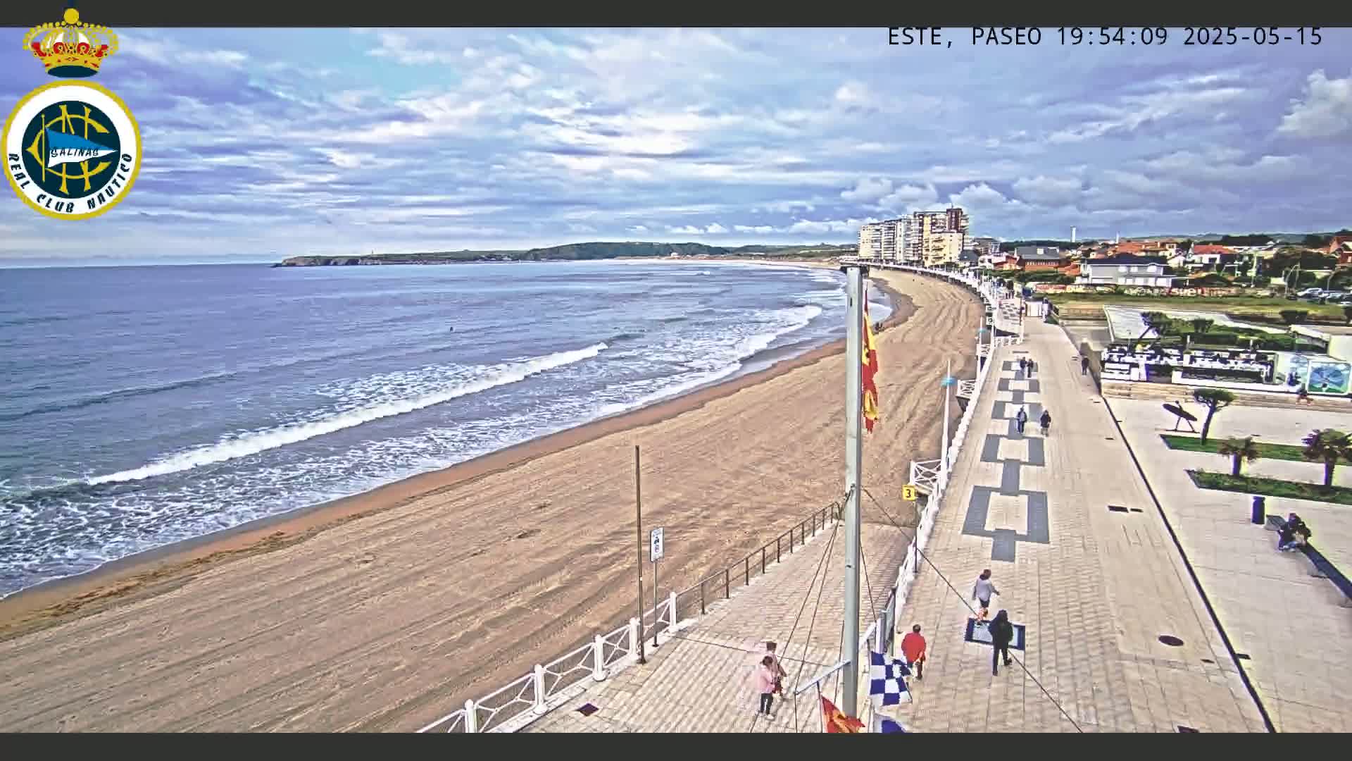 A sandy beach with gentle waves stretches alongside a paved boardwalk, with a few people strolling under a partly cloudy sky.