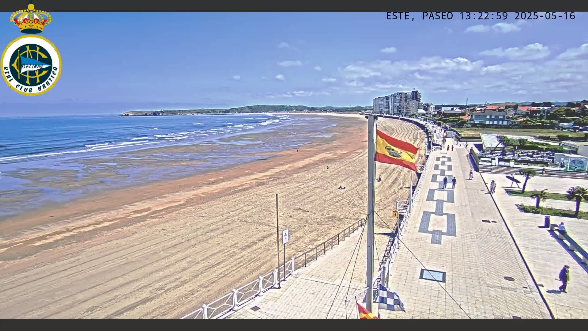 A wide sandy beach with gentle waves under a mostly sunny sky is bordered by a paved walkway with a Spanish flag, and a few people are seen walking along the promenade.