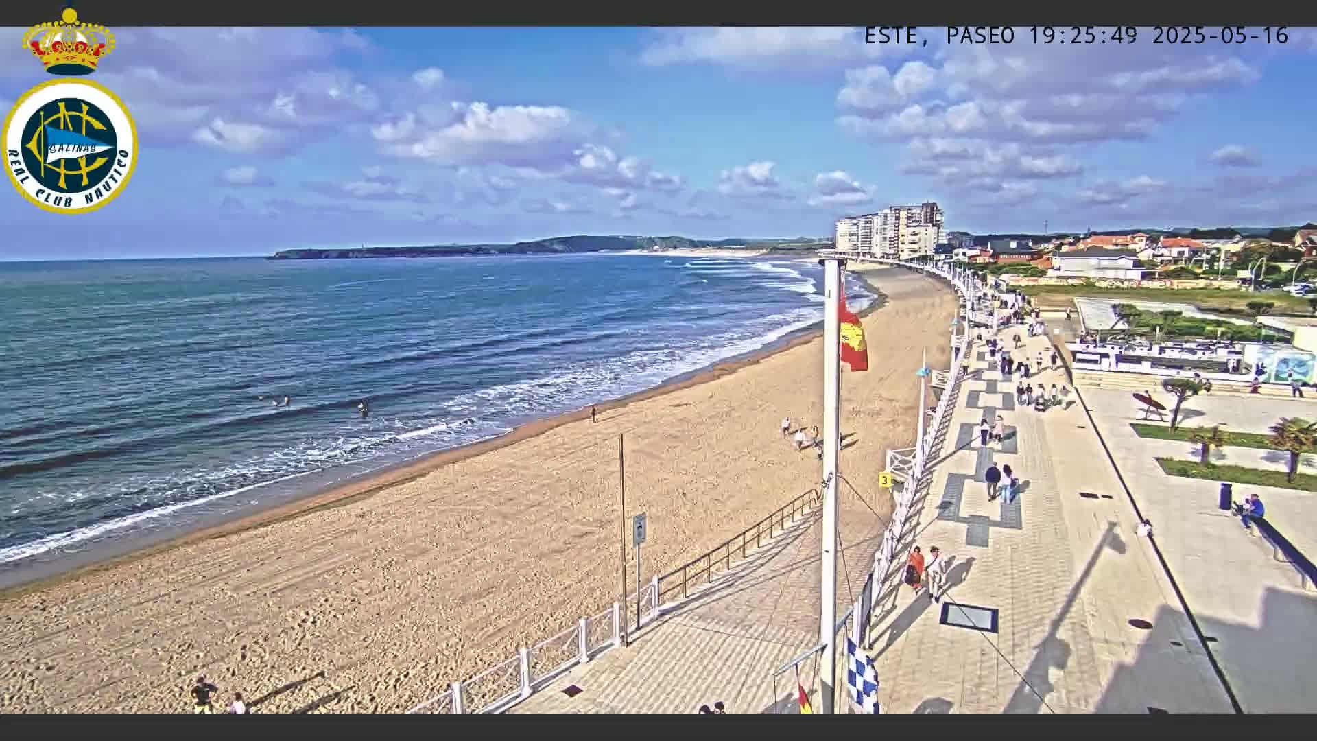 A sandy beach with calm ocean waves and a paved walkway lined with people extends from a shoreline under a partly cloudy sky.