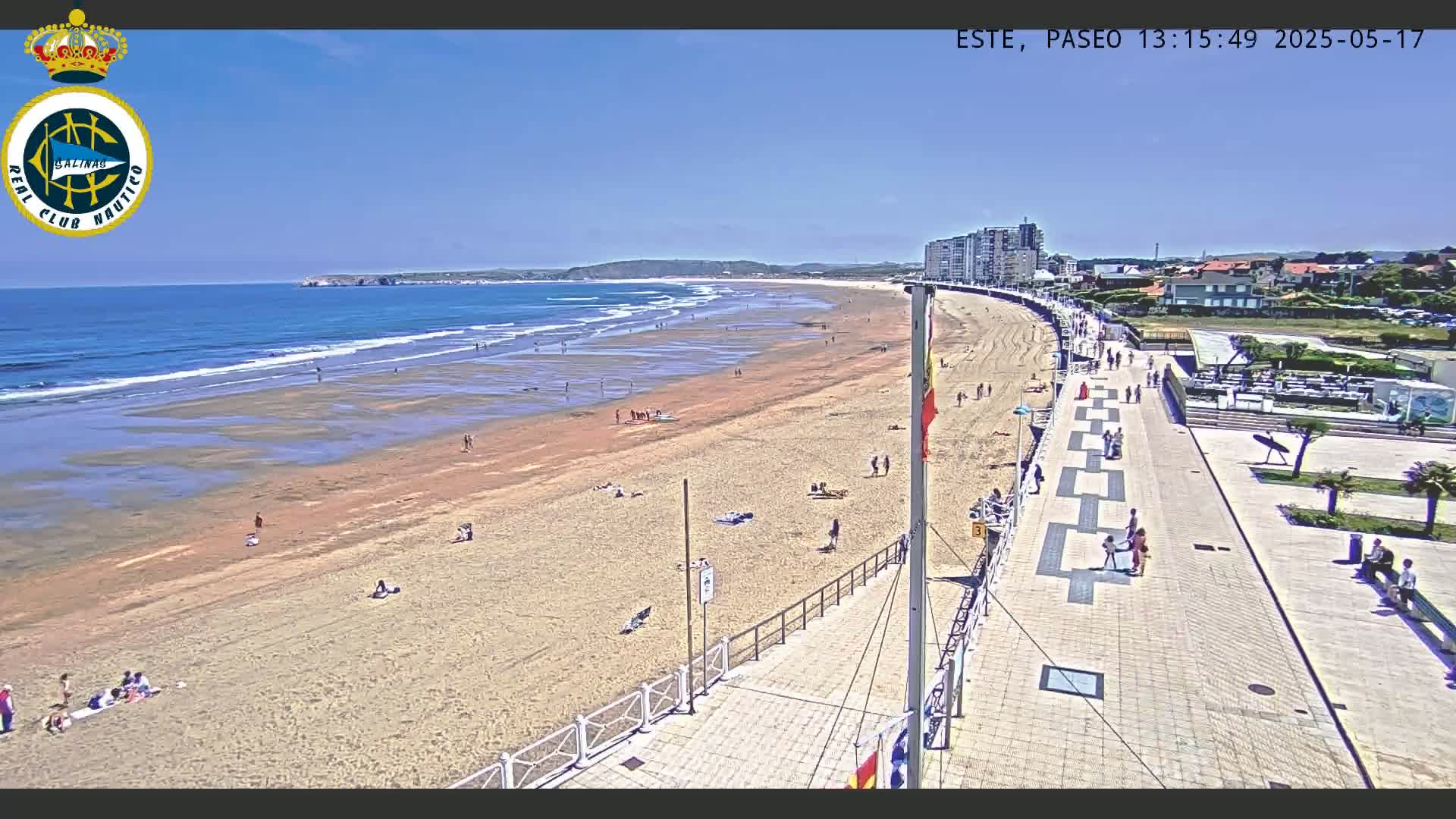 A sandy beach with scattered people, a boardwalk, and buildings in the background under a clear, sunny sky.