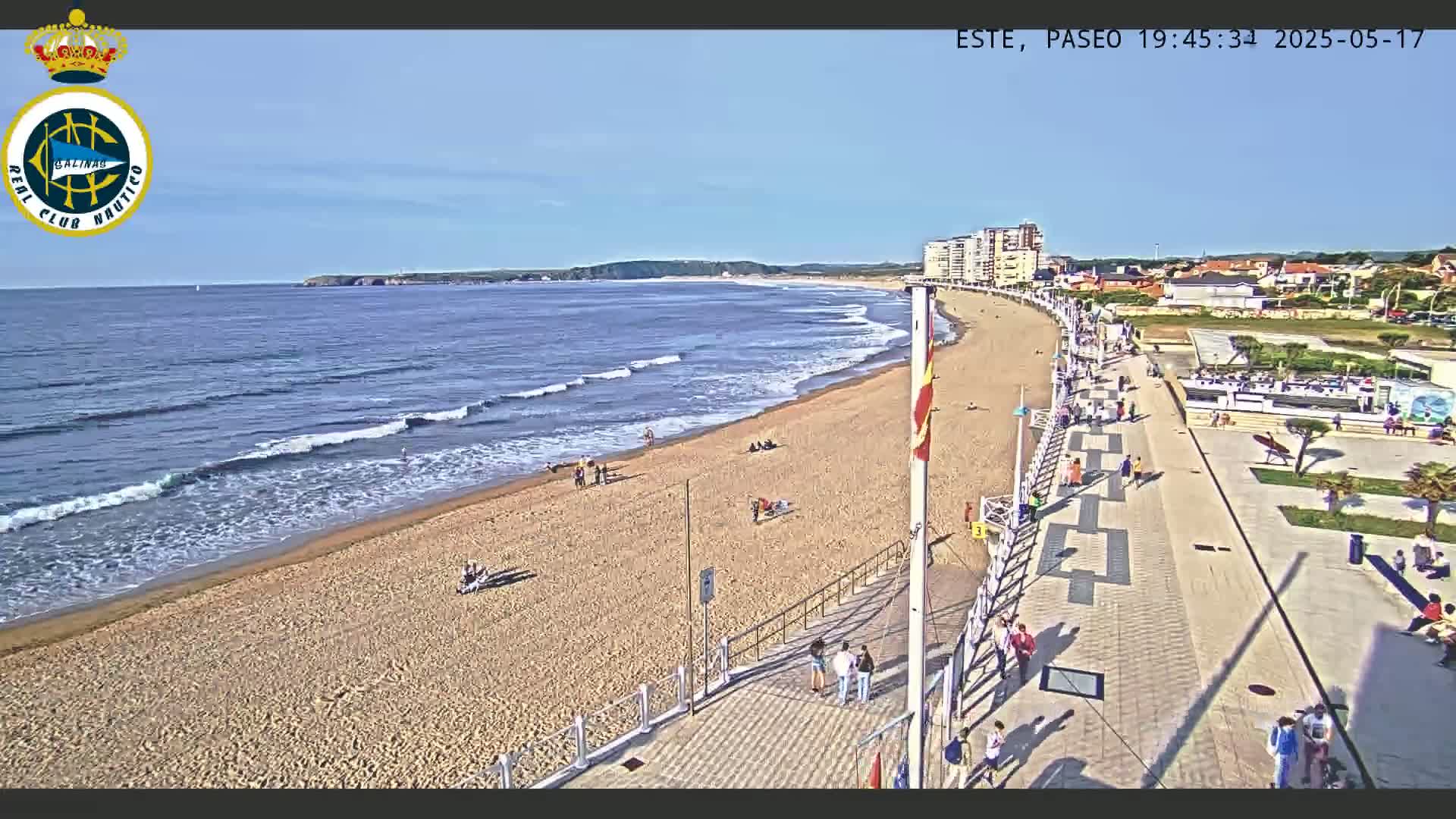 A sandy beach with gentle waves and scattered people is bordered by a paved promenade with buildings and more people along it, all under a clear sunny sky.