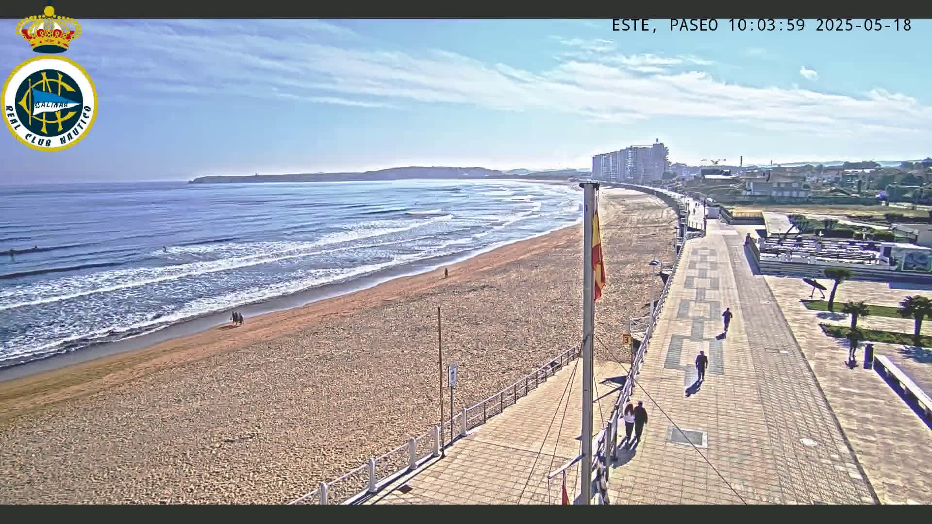A sandy beach with gentle waves under a mostly sunny sky is seen from above, next to a paved boardwalk with a few pedestrians.