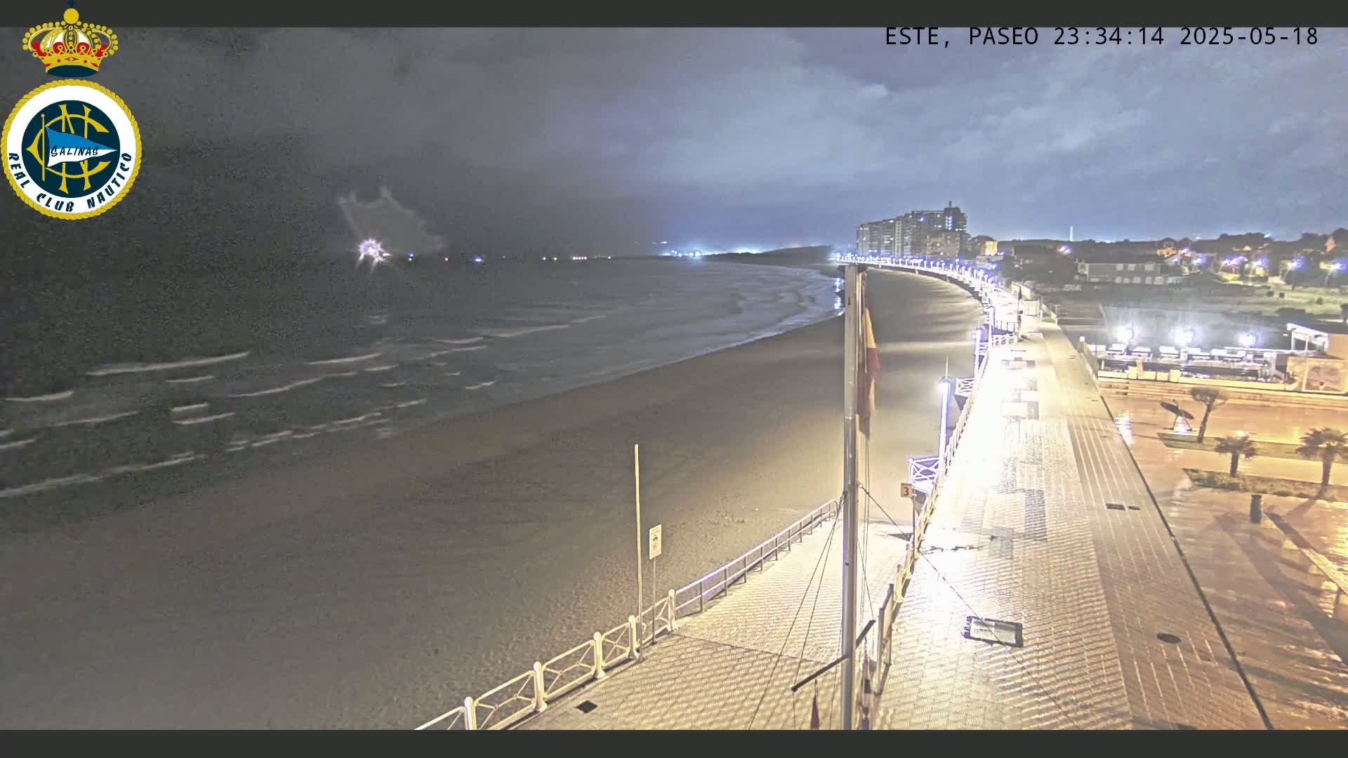 A nighttime beach scene shows a dark, wavy ocean under an overcast sky, with a paved boardwalk and buildings lit up in the background.