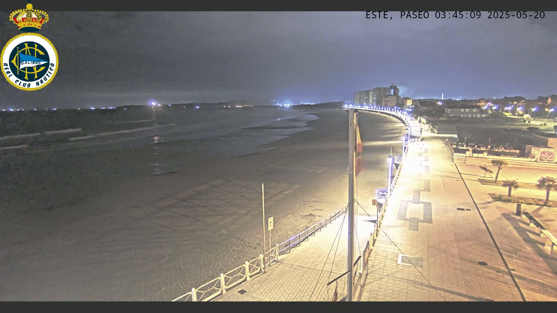 A nighttime, high-angle view of a mostly empty beach and boardwalk with buildings and lights visible in the distance.