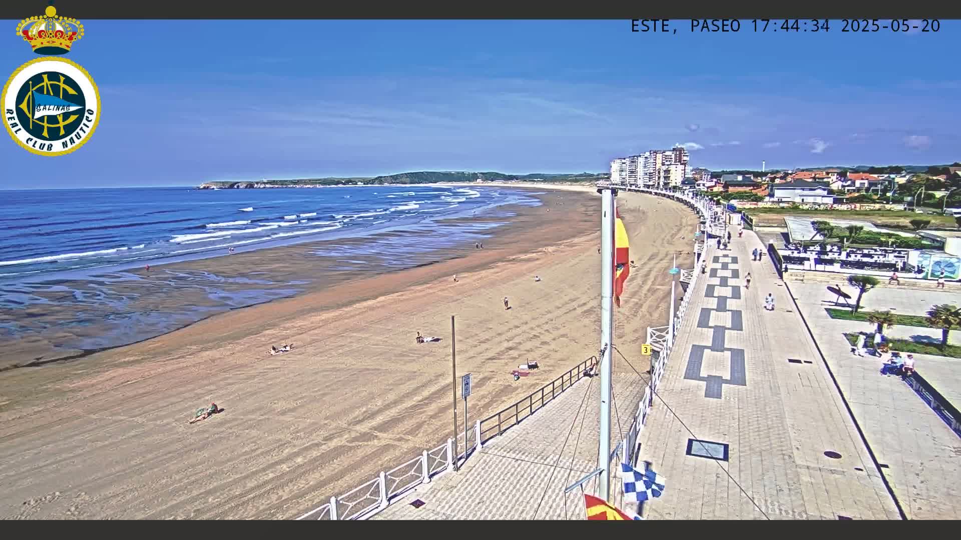 A sandy beach with moderate waves under a clear blue sky is bordered by a paved walkway and lined with buildings.