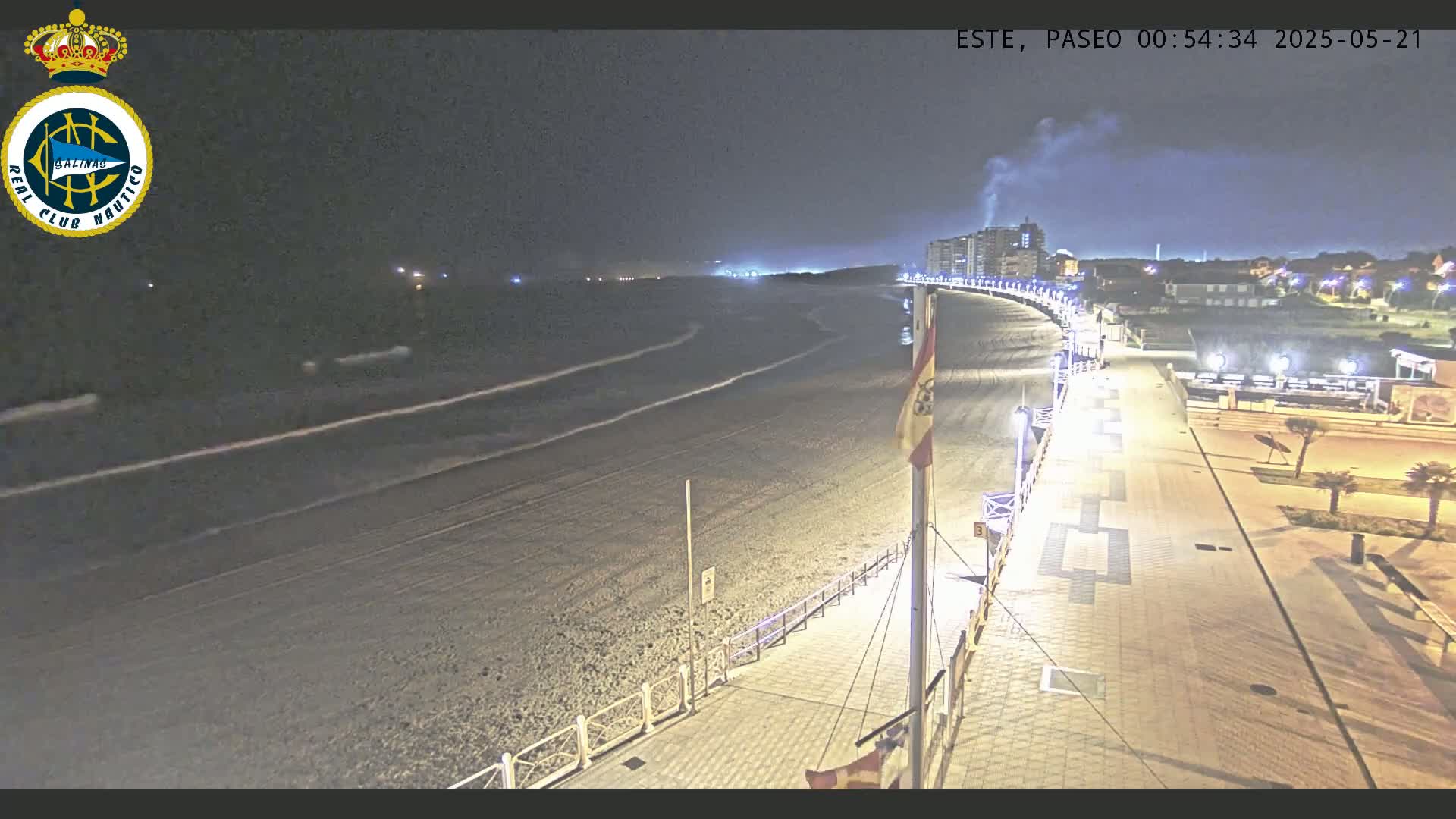 A nighttime, high-angle view shows a mostly empty, dark sandy beach, a paved boardwalk with lights, and buildings in the distance under a clear sky.