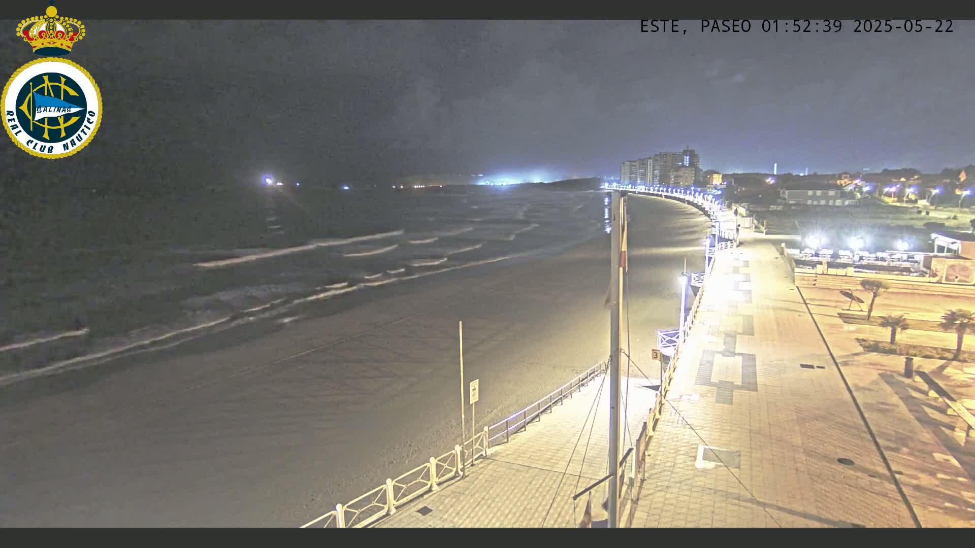 A nighttime, high-angle view shows a sandy beach with gentle waves lapping the shore, a paved boardwalk lined with lights, and buildings in the distance under a dark sky.