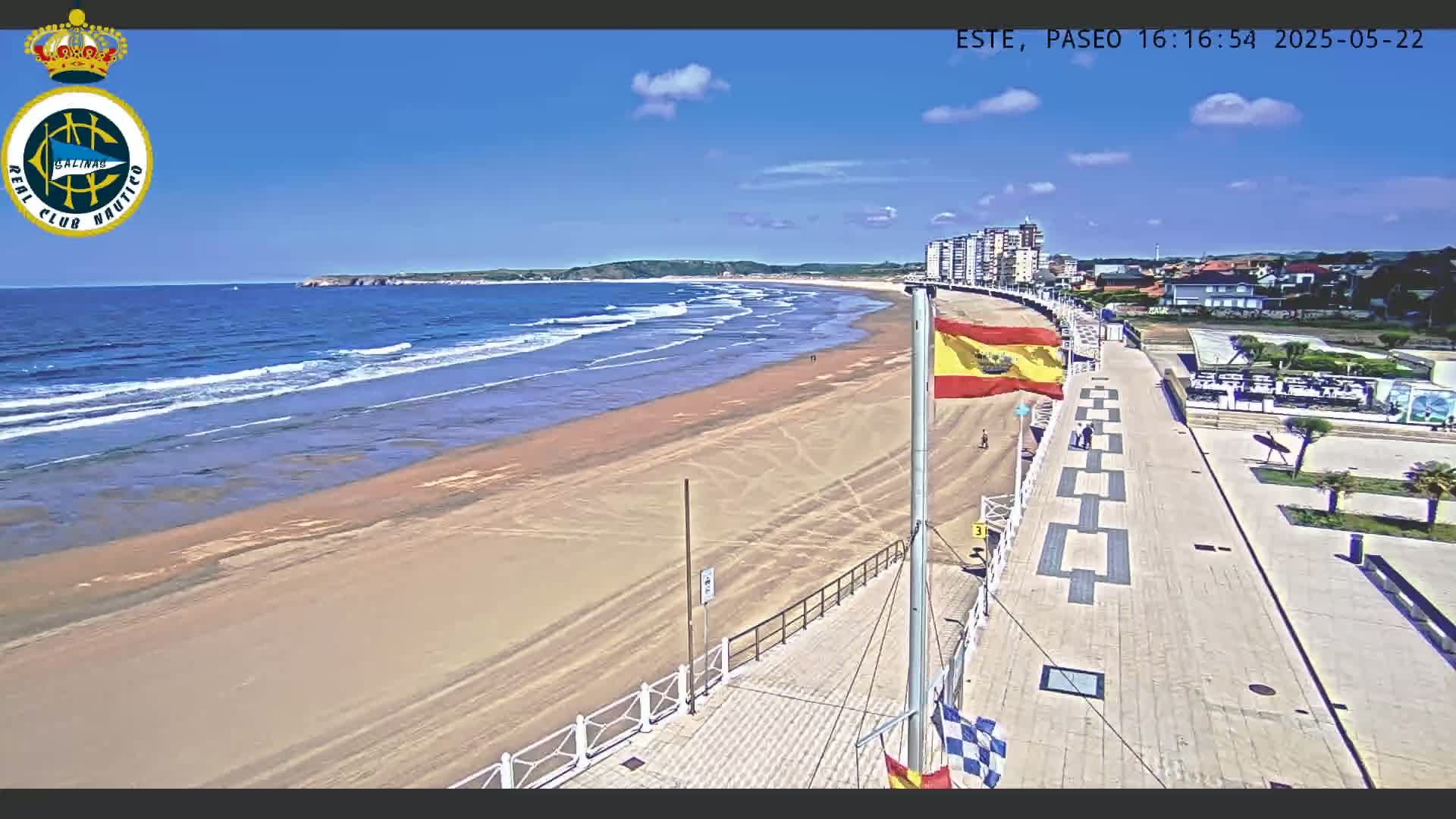 A sandy beach with gentle waves under a sunny sky is bordered by a paved boardwalk and buildings.