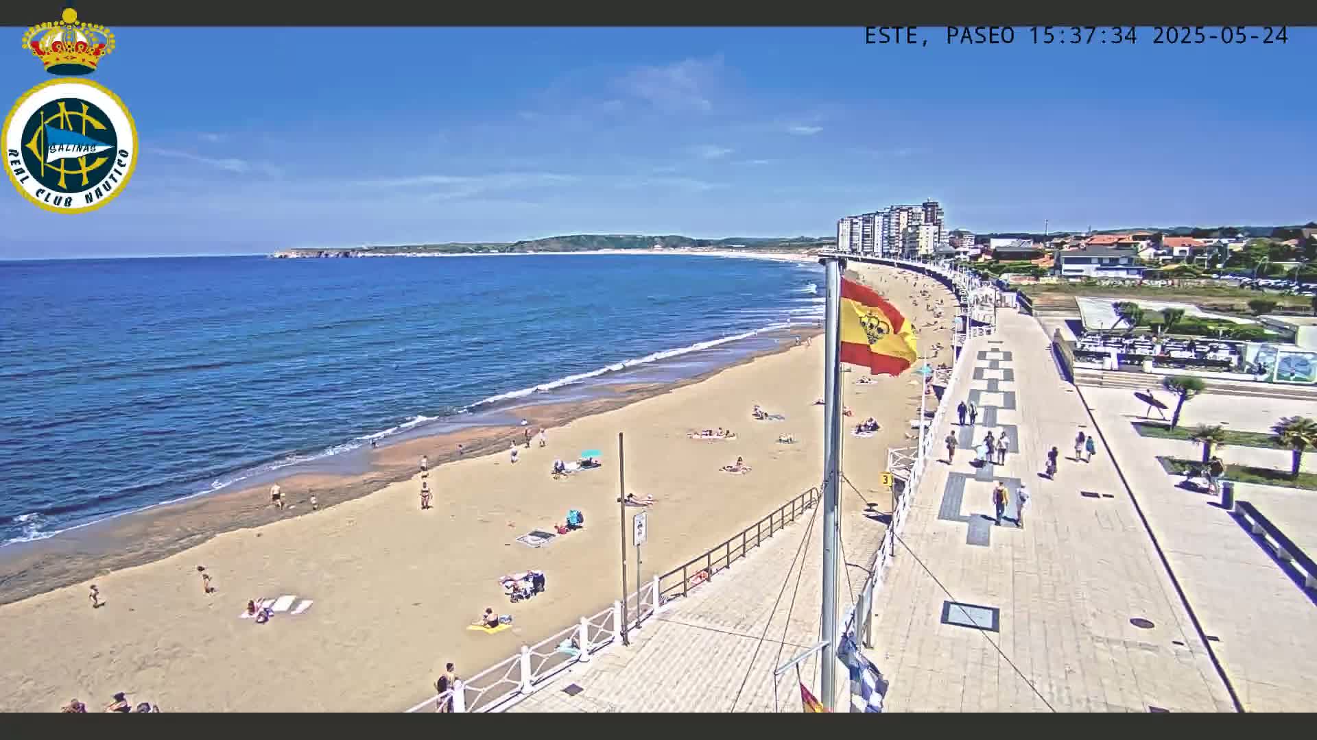 A sunny beach with a sandy shoreline and a boardwalk lined with people extends to a calm ocean under a clear blue sky.