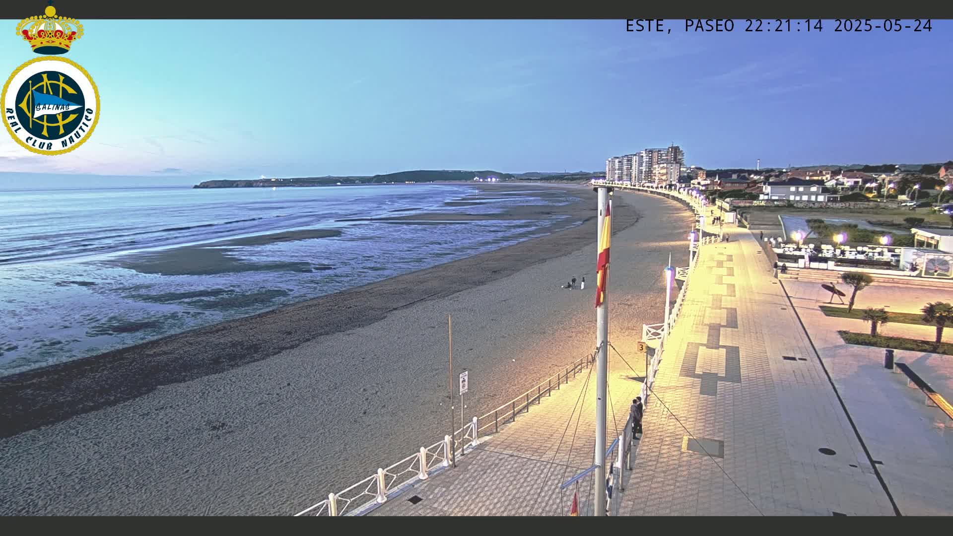 A long, sandy beach at twilight is bordered by a paved walkway lined with lights, leading to buildings and a calm ocean.
