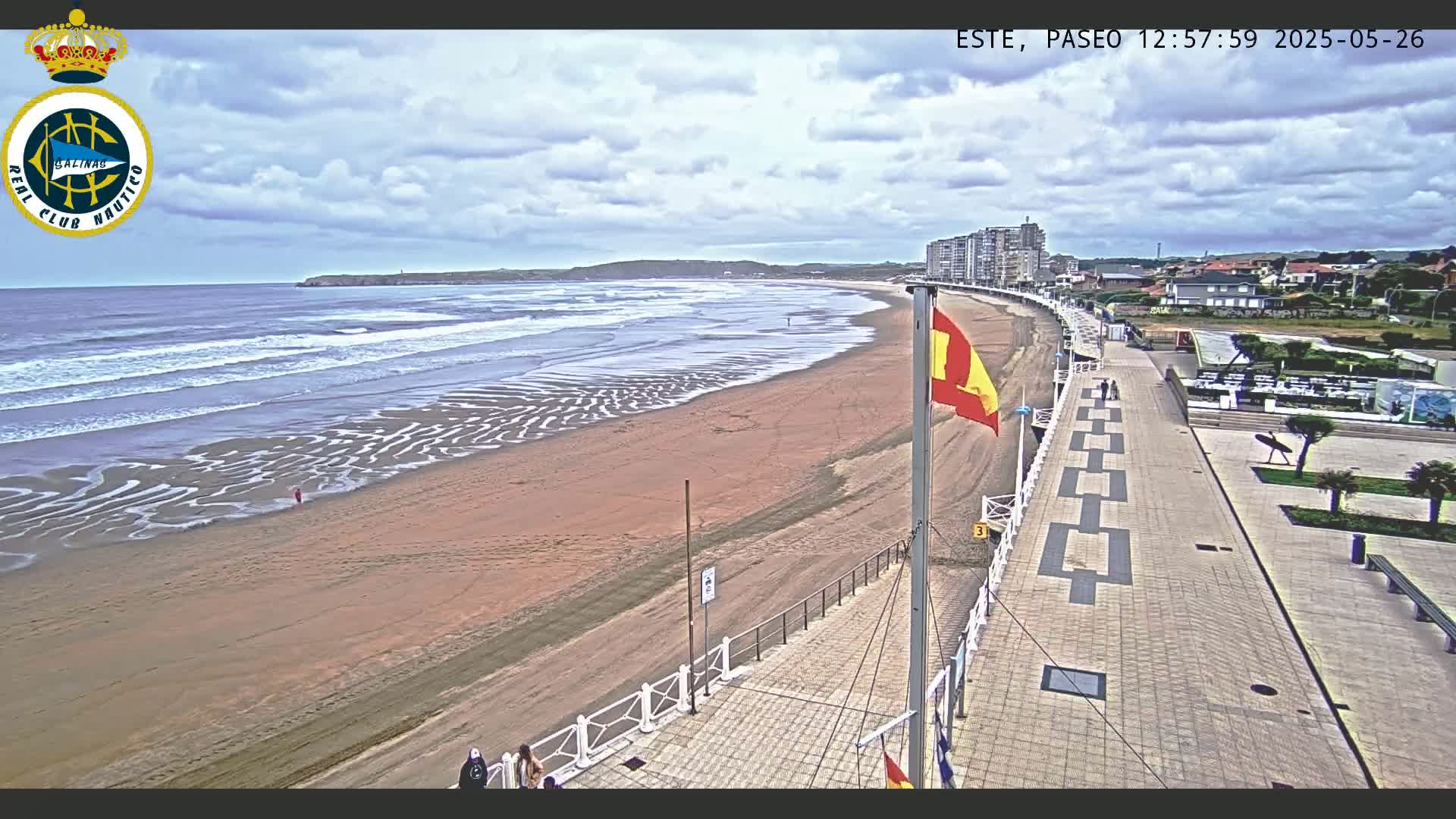 A sandy beach with moderate waves under a partly cloudy sky is adjacent to a paved boardwalk lined with buildings.