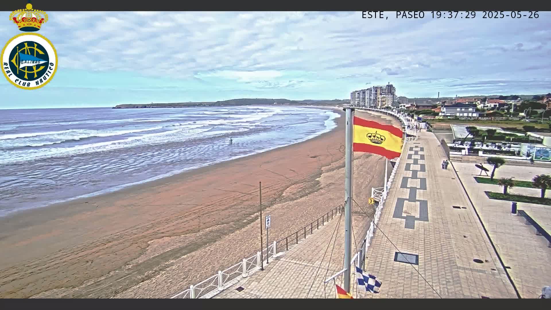 A sandy beach with gentle waves under a partly cloudy sky is overlooked by a paved boardwalk and buildings.